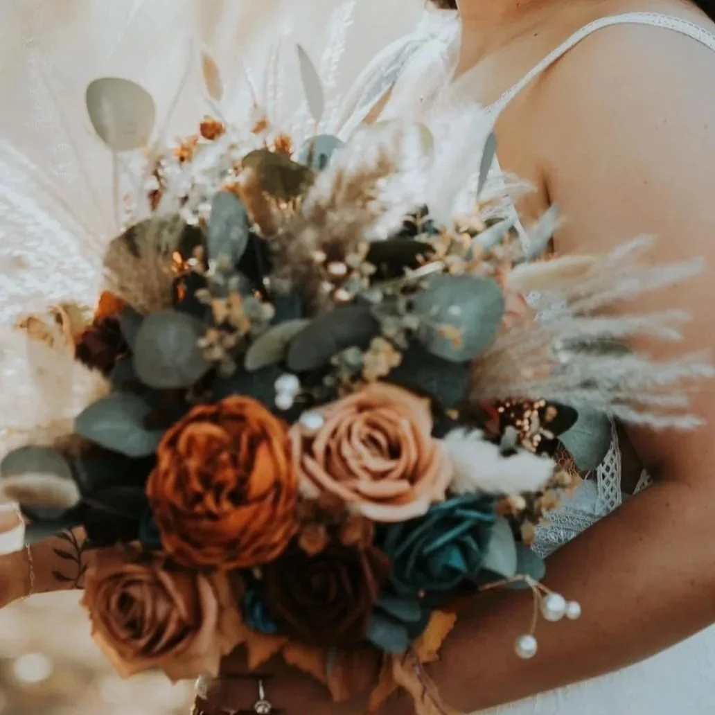 Person holding a bouquet of flowers, including roses and eucalyptus, with decorative feathers.