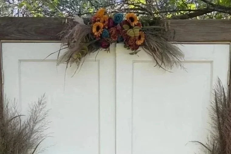 Decorative floral arrangement on top of white double doors with wooden frames, surrounded by dried grasses.