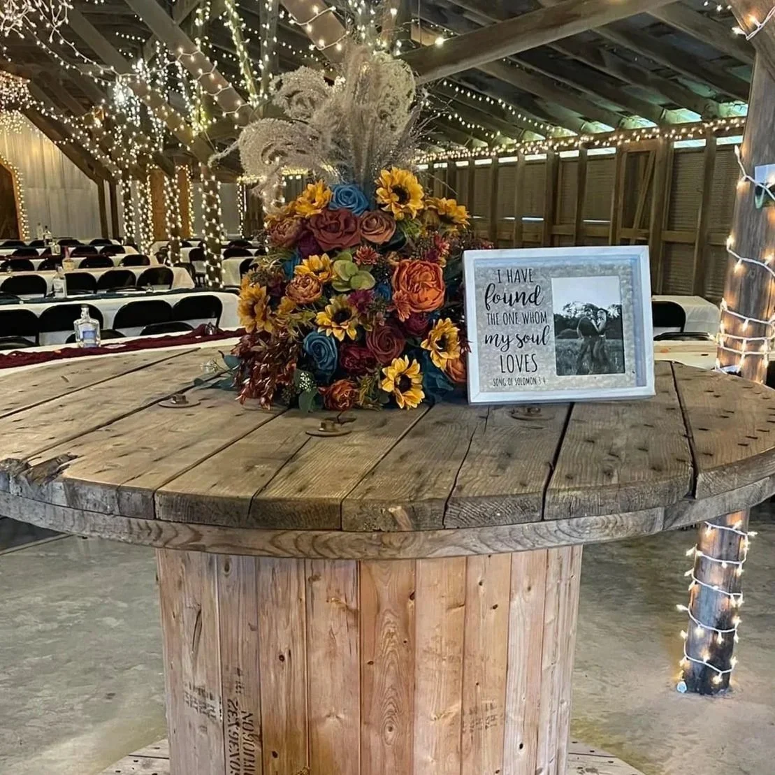 A floral arrangement with roses, sunflowers, and decorative grasses on a rustic wooden table. A framed sign with a quote and a black and white photo is beside the arrangement. The background features a barn-like setting with string lights and tables,