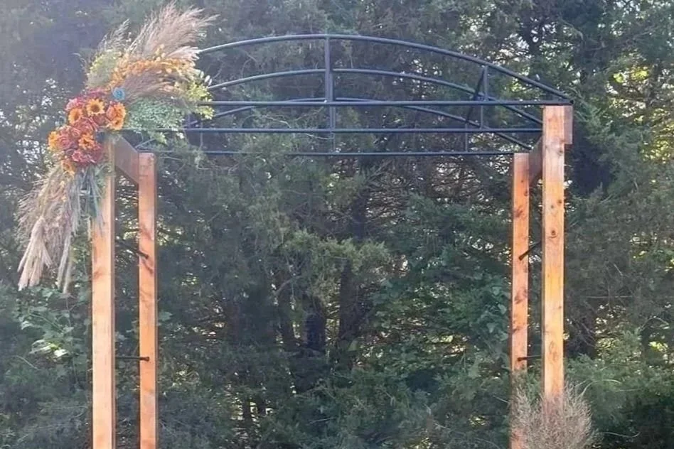 Wooden archway decorated with a bouquet of flowers and grasses, set outdoors with trees in the background.