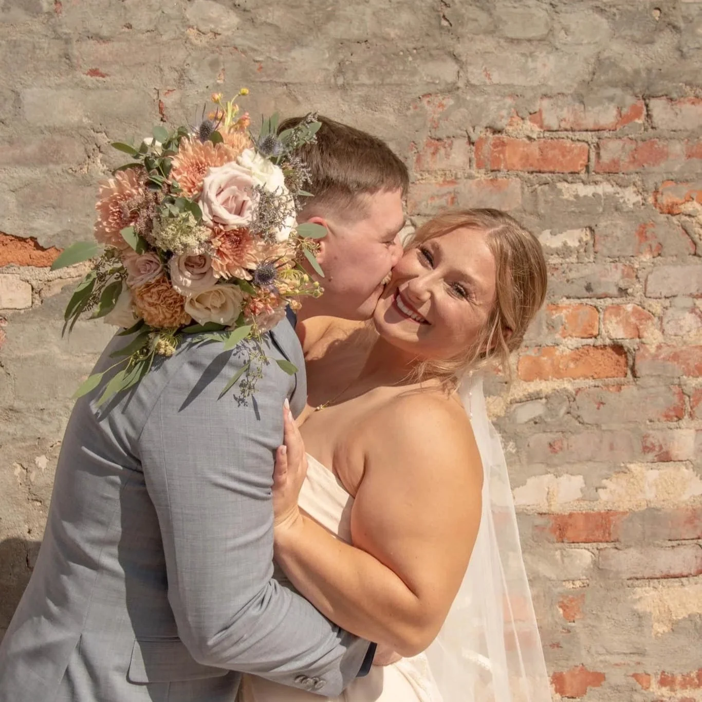 A bride and groom are happy and embracing on their wedding day, with the groom giving the bride a kiss on the cheek. The groom is holding a bouquet of flowers, and they are standing against a brick wall.