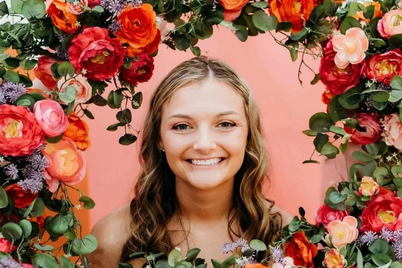 A bride smiling surrounded by a decorative floral arch with pink, red, orange, and purple flowers and green leaves, set against a pink background.