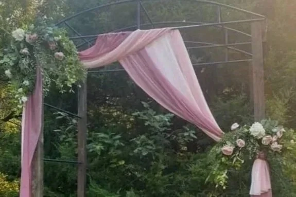 Outdoor wedding arch decorated with pink and white fabric and floral arrangements, set against a natural green background.