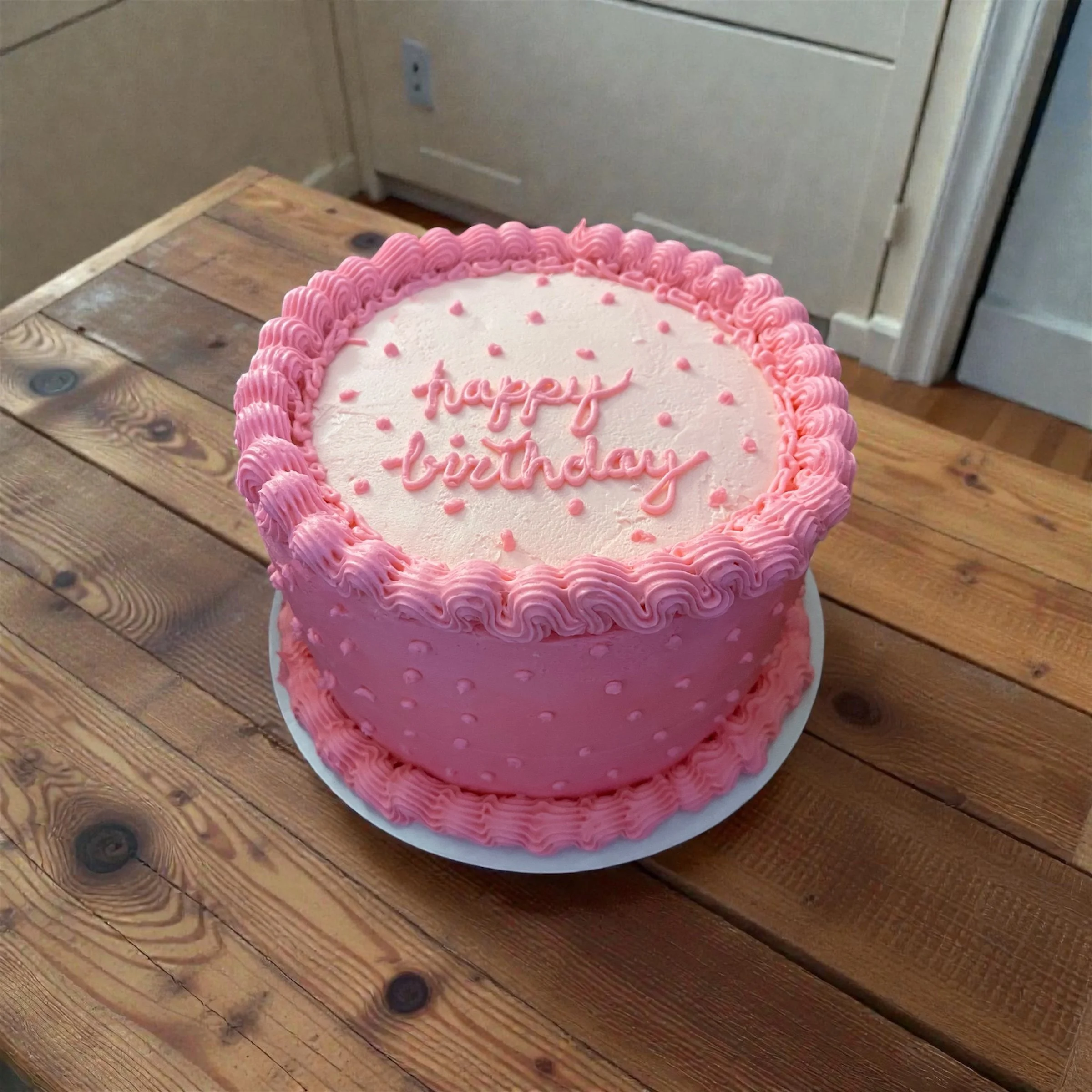 A pink birthday cake with decorative icing on the edges and small pink dots on the sides. The top has a message in pink icing that says "happy birthday" with pink dots scattered around.