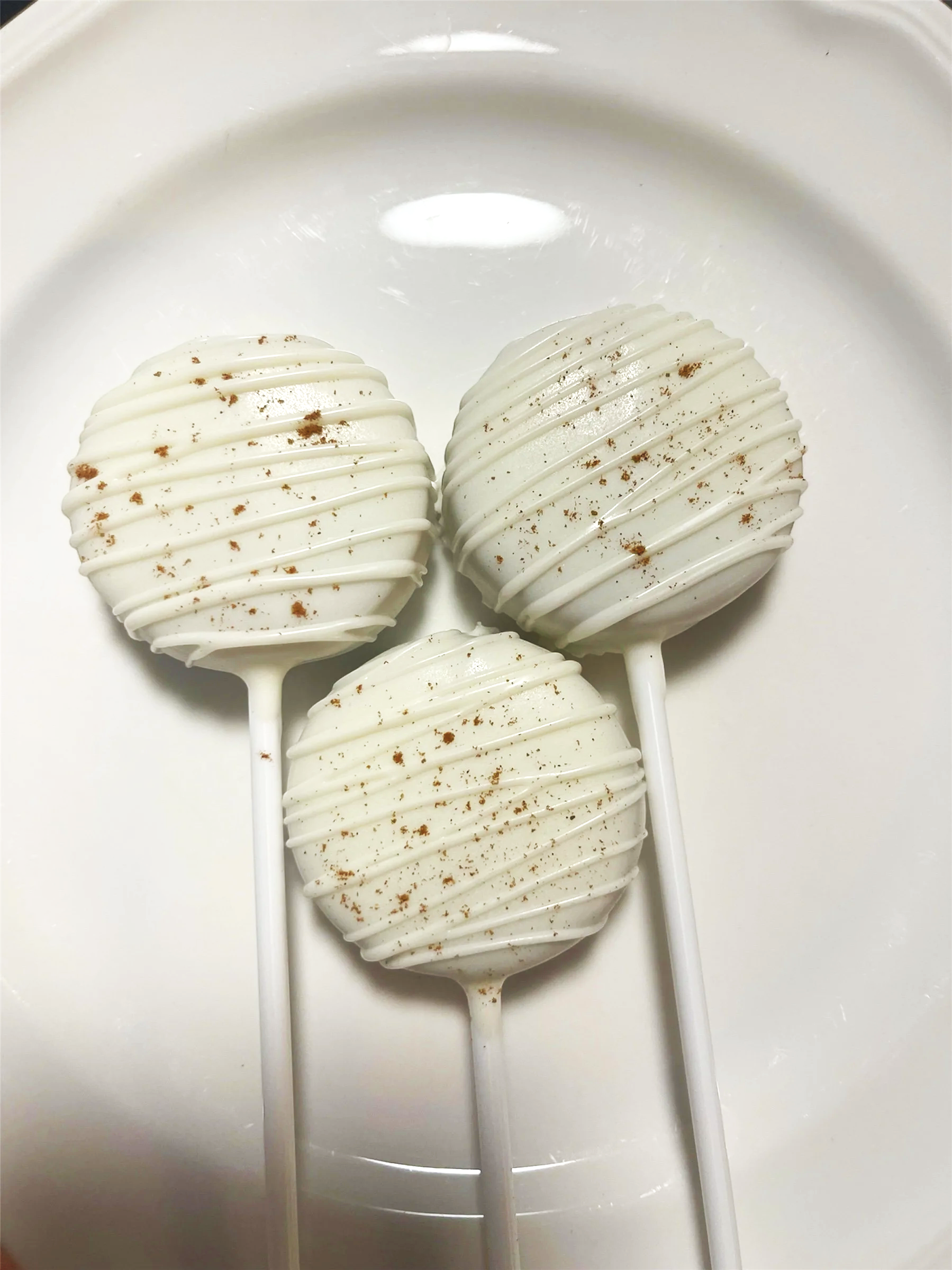 Three white cake pops decorated with white chocolate drizzle and sprinkled with brown crumbs, placed on a white plate.
