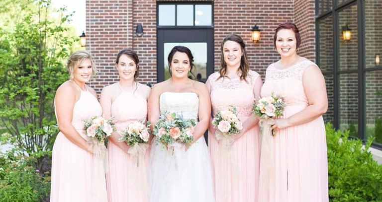 Five women in pastel pink and white dresses standing outdoors in front of a brick building, holding bouquets of flowers.