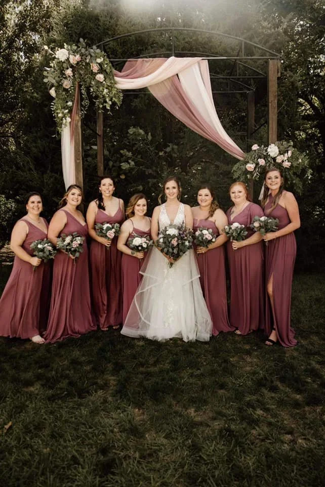 Group of eight women dressed in mauve bridesmaid dresses, standing outdoors in front of a wooden arch decorated with white and pink drapery and floral arrangements, during a wedding celebration.