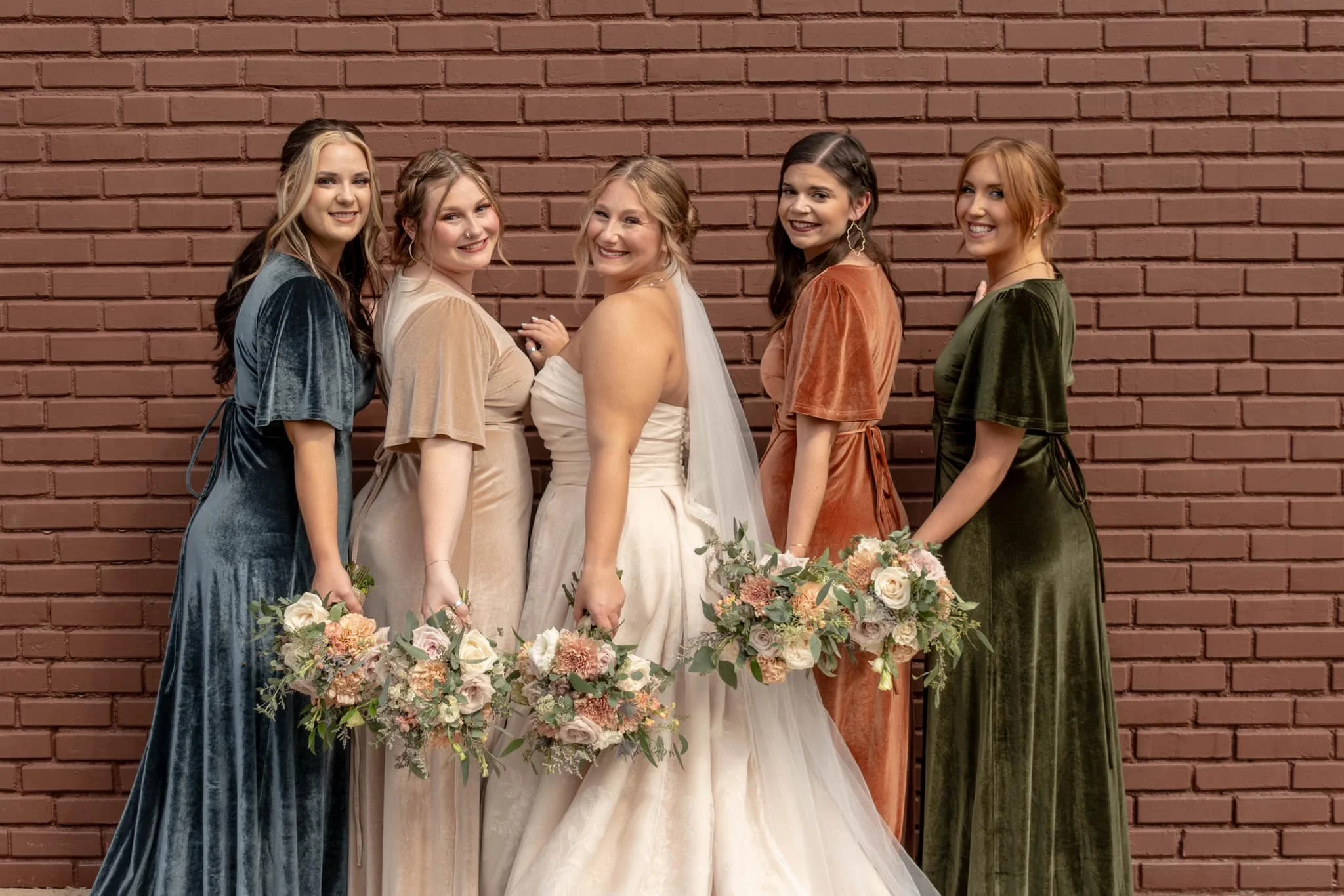 Six women standing in a row against a brick wall, holding bouquets of flowers, with one woman wearing a wedding dress and veil in the center, celebrating a wedding.