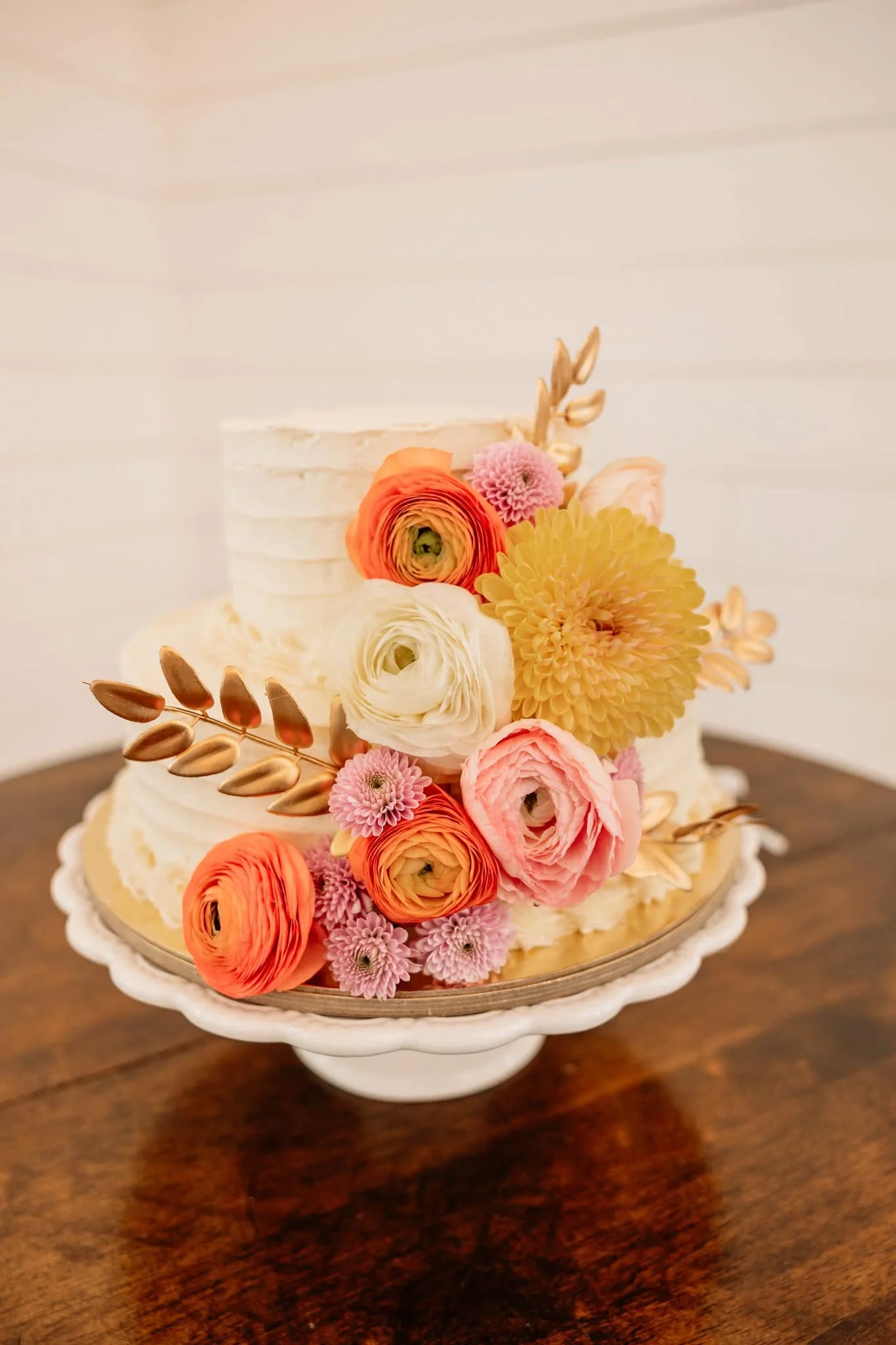 A two-tiered white wedding cake decorated with pastel-colored flowers, including ranunculus, chrysanthemums, and dahlias, on a white cake stand placed on a wooden table.
