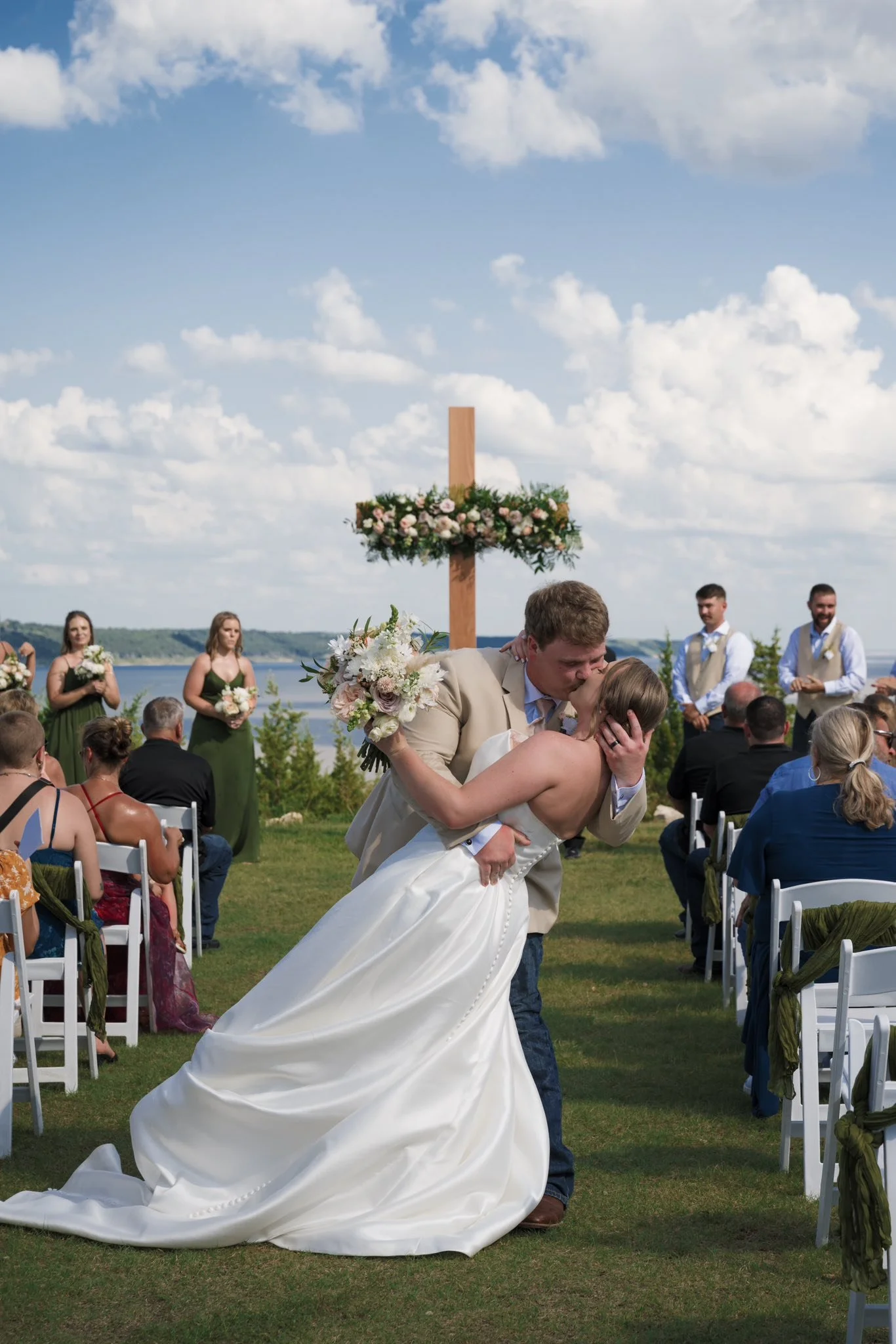 A couple dressed in wedding attire sharing a kiss during an outdoor wedding ceremony near a river, with guests seated and standing around.