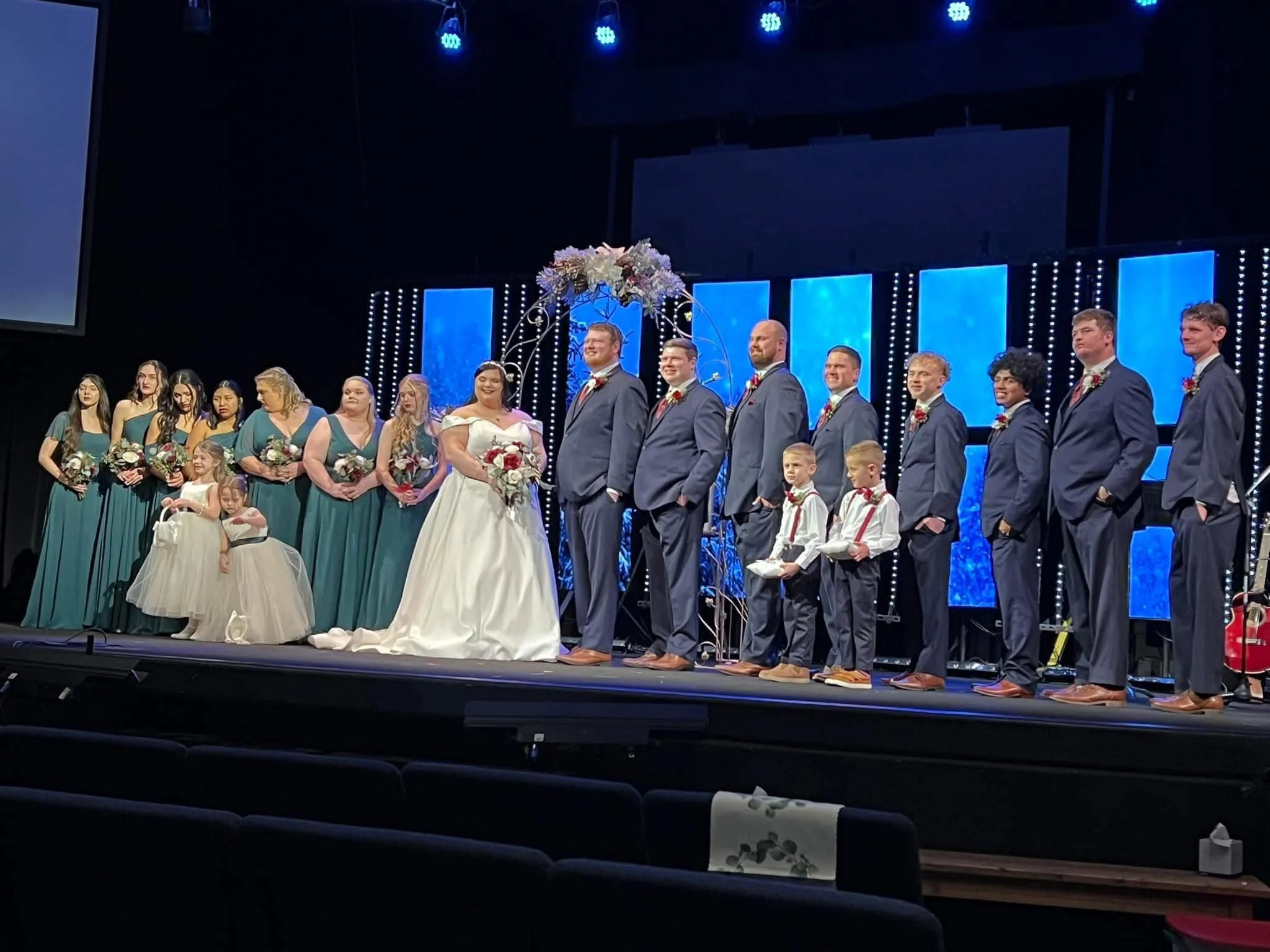 A wedding ceremony taking place on stage with the bride and groom at the center, surrounded by bridesmaids, flower girls, groomsmen, and ring bearers. The stage has a decorated arch with flowers and lights in the background.