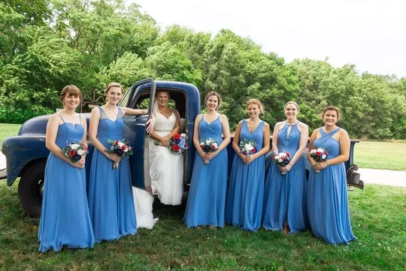 A bride in a white dress standing in a vintage blue truck surrounded by six bridesmaids in matching blue dresses holding bouquets, outdoors on a grassy area with trees in the background.