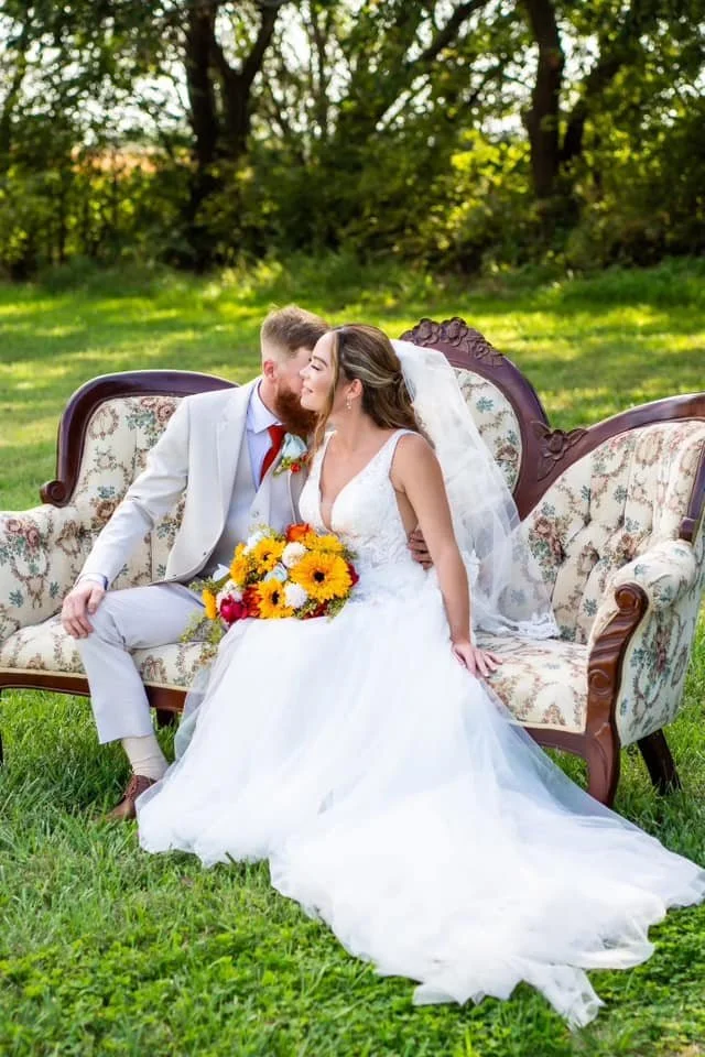 A bride and groom sitting close together on a vintage floral sofa outdoors during a wedding photoshoot, the bride holding a colorful bouquet, surrounded by green grass and trees.