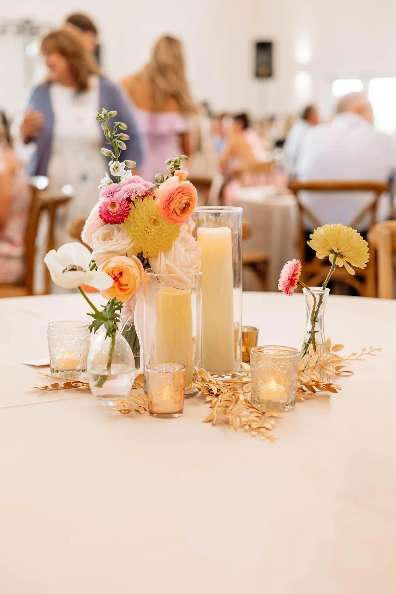 A decorated table with pink, white, and peach flowers in vases, surrounded by candles in glass holders, at what appears to be a wedding or special event reception.