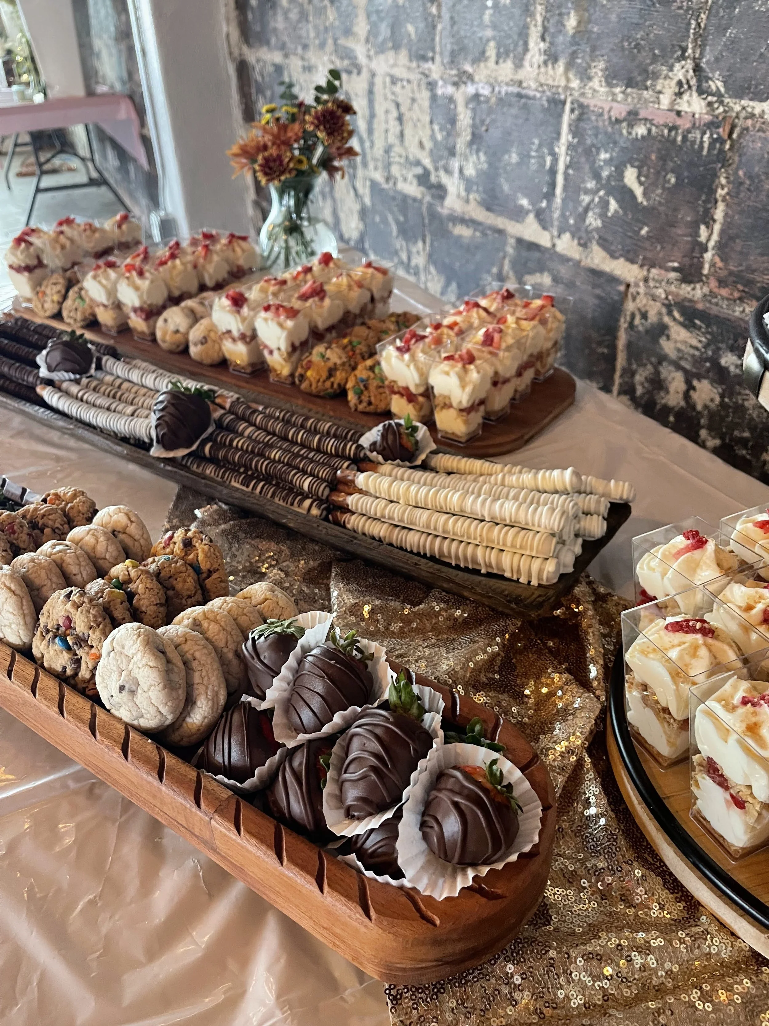 A dessert table with assorted sweets including cookies, chocolate-covered strawberries, layered pudding cups with fruit, and decorated bars, on a table with a gold sequined tablecloth and a brick wall in the background.