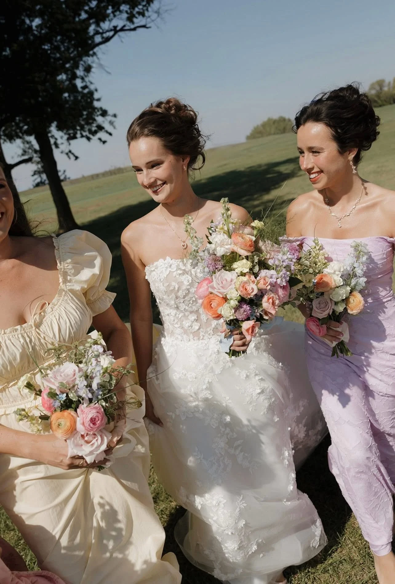 Three women in wedding dresses and pastel-colored outfits standing outside on a grassy field, holding bouquets of flowers, with trees and blue sky in the background.