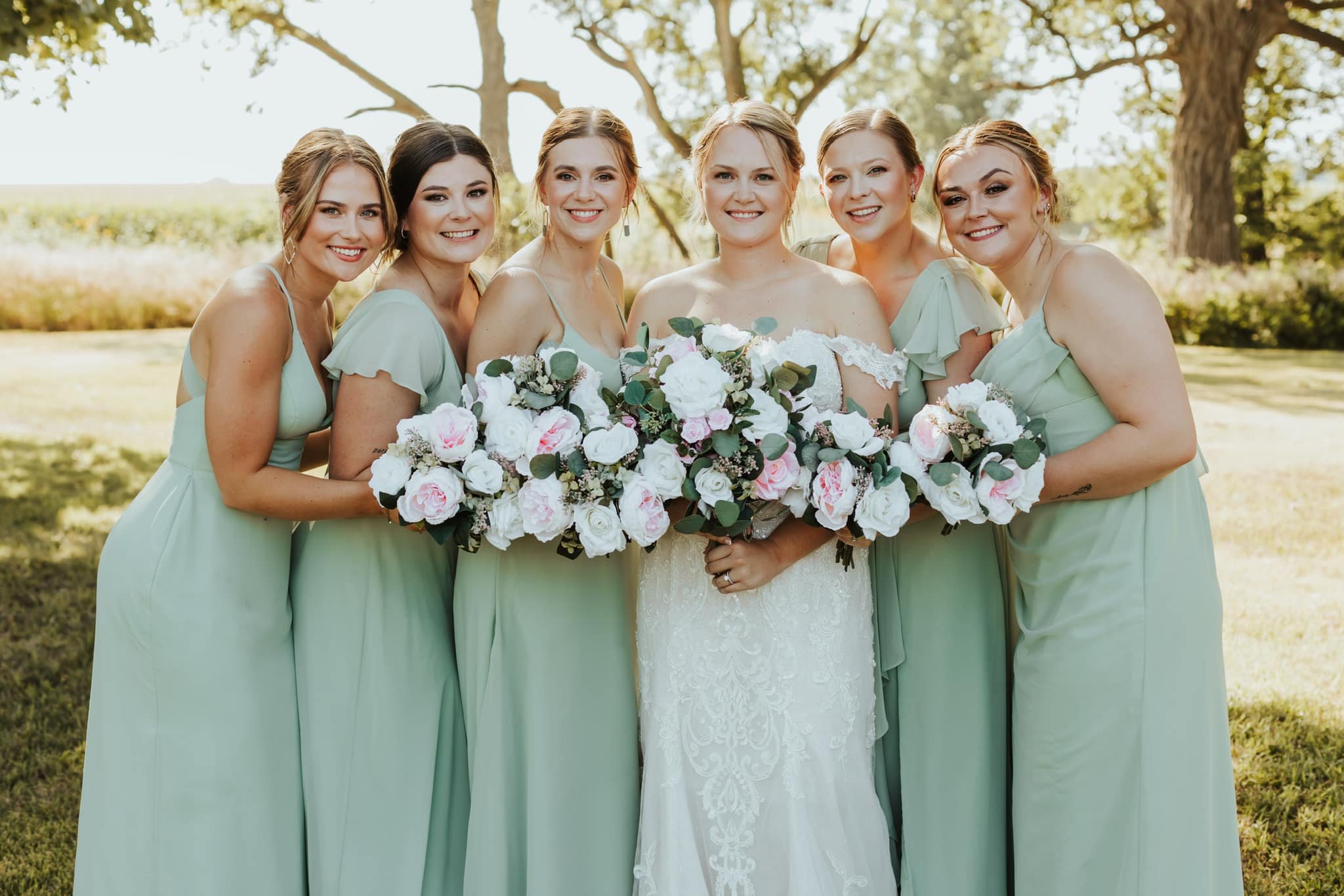 A bride and six bridesmaids in light green dresses standing outdoors, holding floral bouquets of pink and white roses with greenery in a bright, sunny setting with trees in the background.