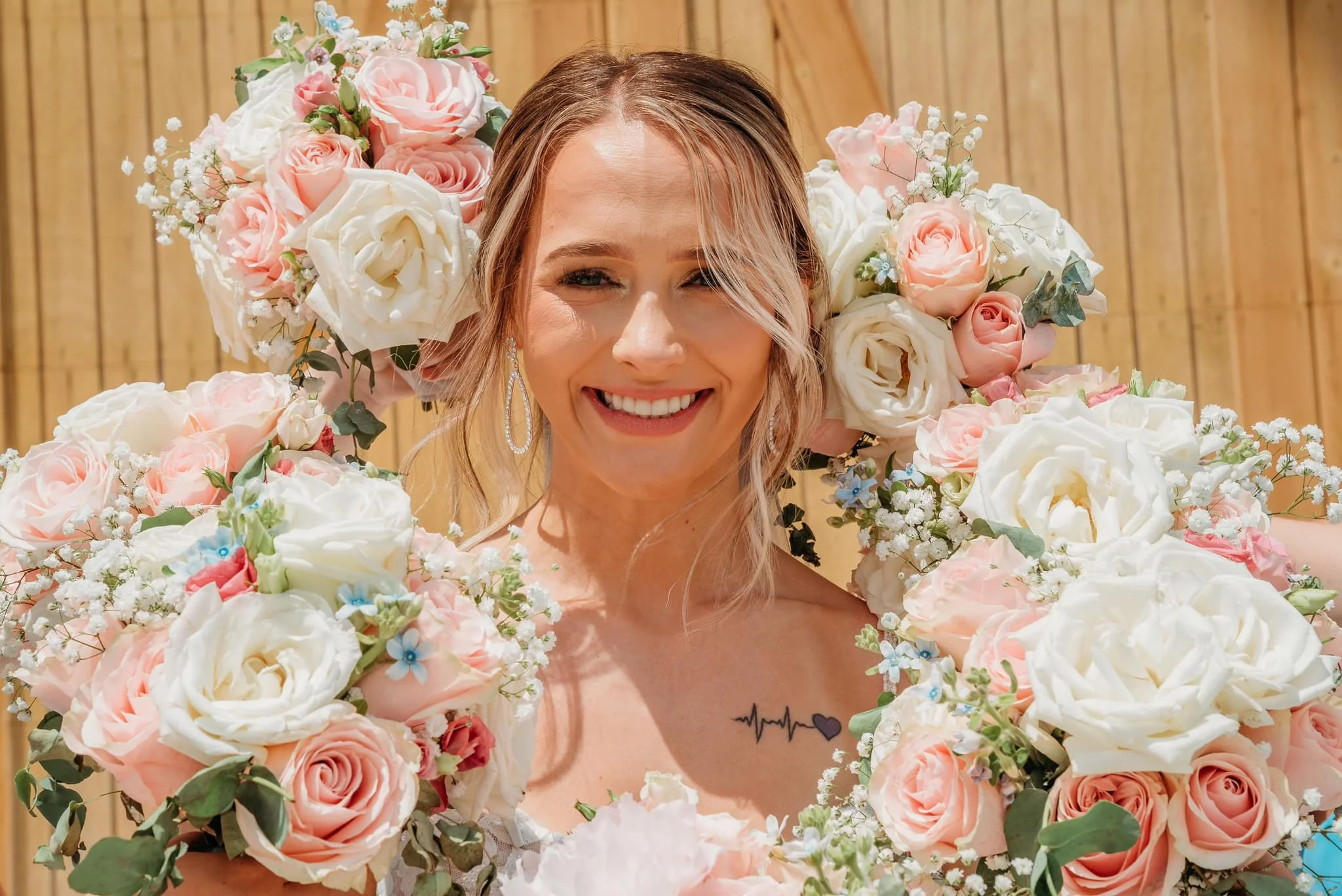 A smiling woman with blonde hair and earrings, surrounded by a large wreath of pink and white roses and small white flowers, standing in front of a wooden fence.