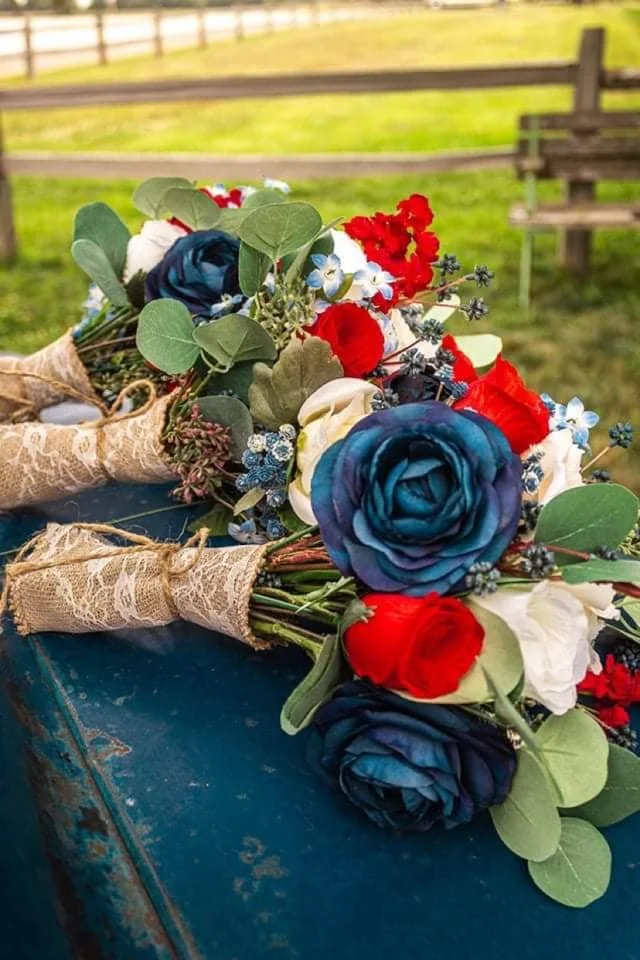 Two bouquets of artificial flowers with blue roses, red and white flowers, and greenery, wrapped in lace, on a blue surface outdoors with a grassy field and wooden fence in the background.