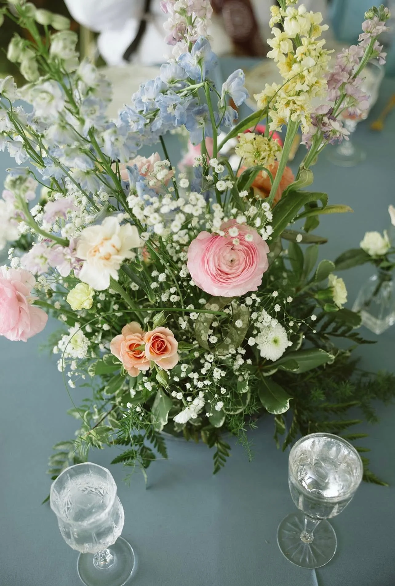 A floral centerpiece with pink, white, yellow, and blue flowers on a table with two empty glasses.