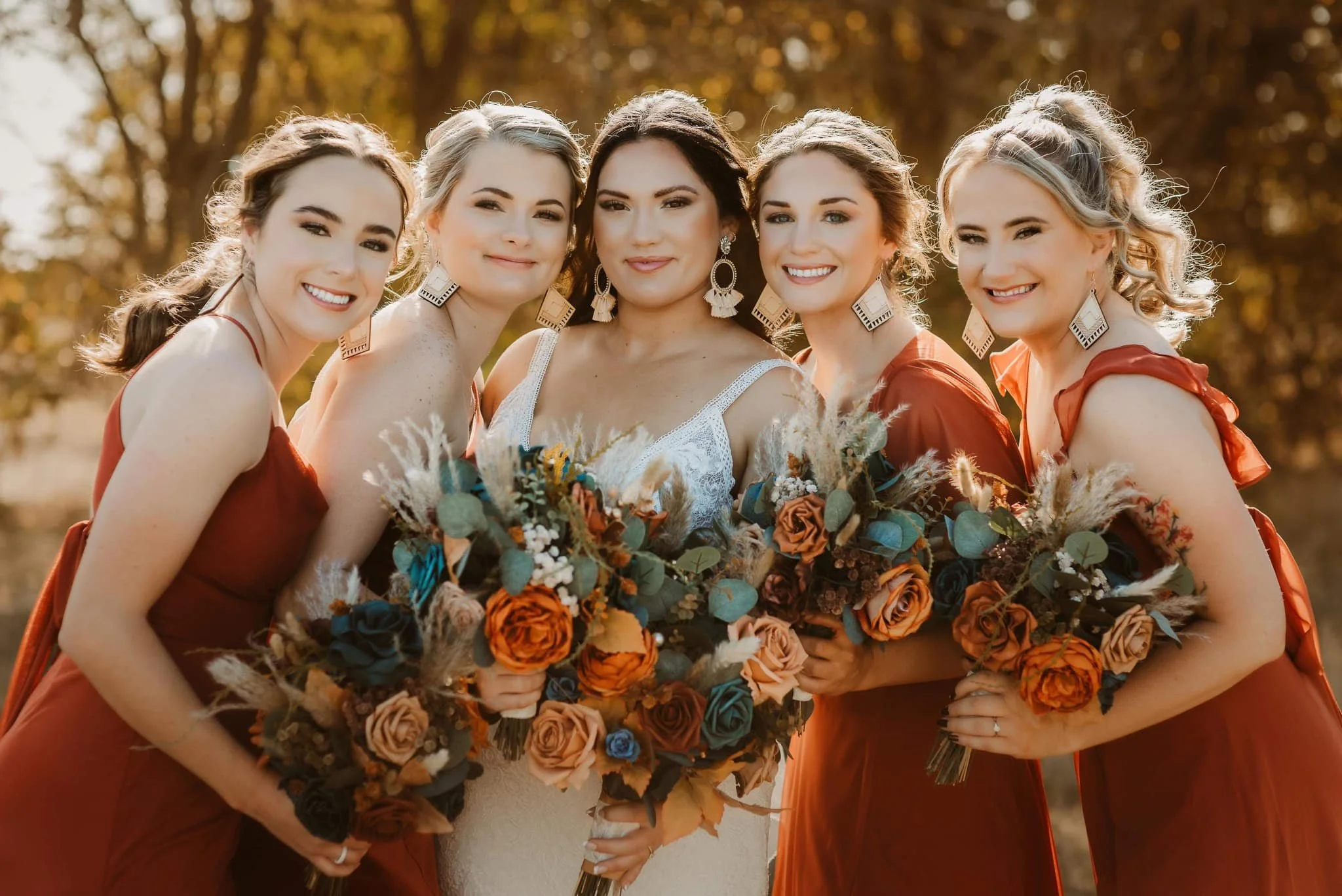Group of five women in fall-colored dresses holding large bouquets, standing outdoors with trees in the background.