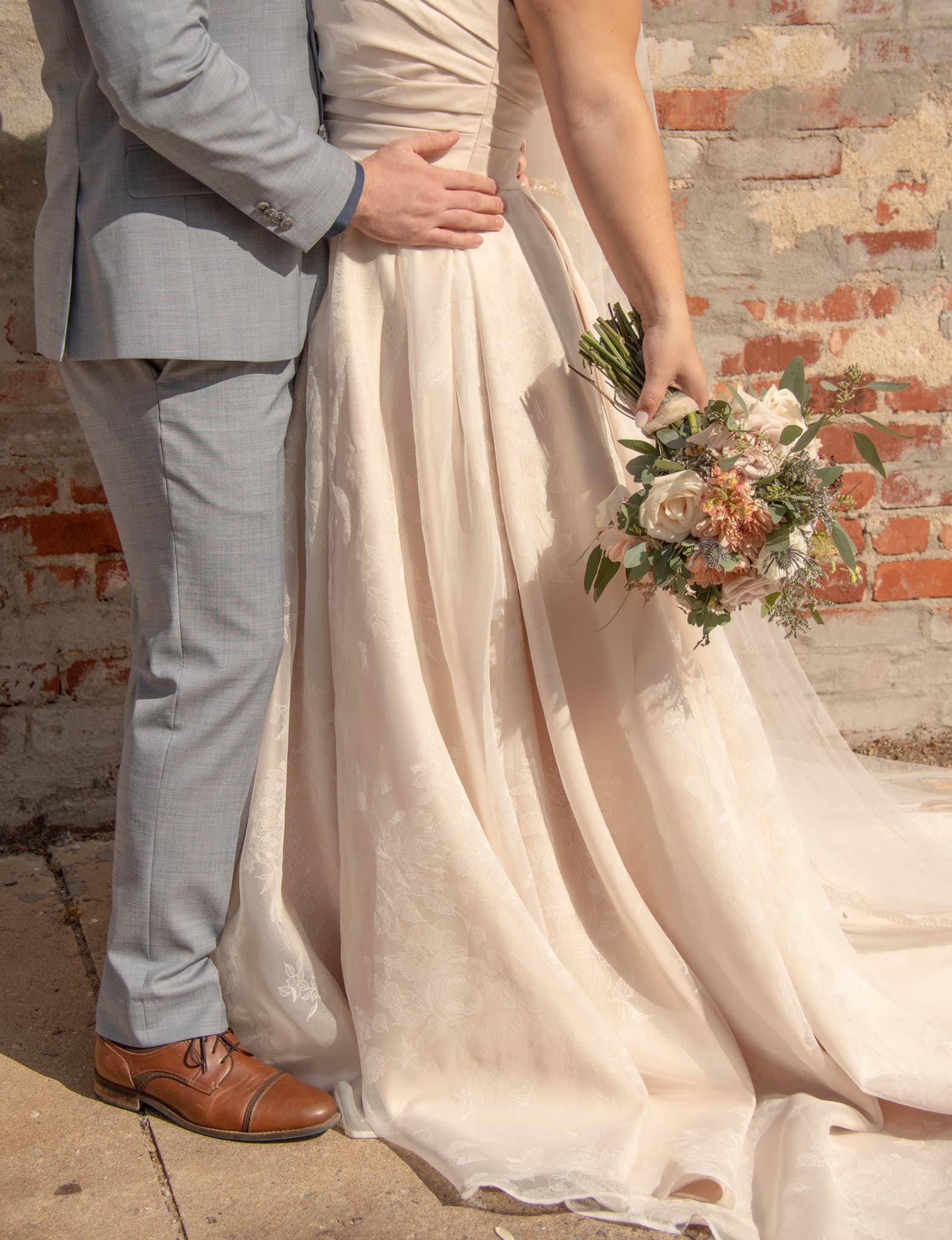 Close-up of a bride and groom standing together, focusing on their torsos and lower bodies. The bride is holding a bouquet of flowers, and they are standing in front of a brick wall.