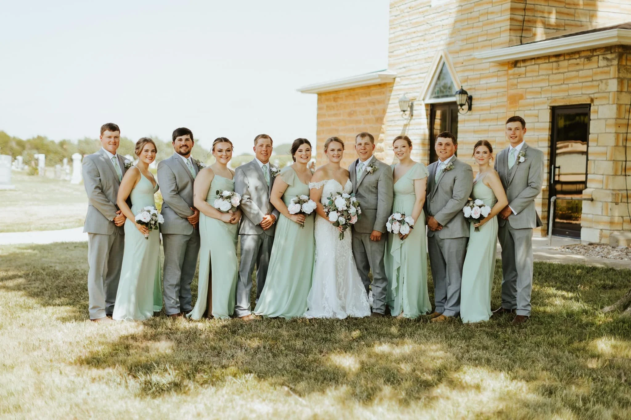 A wedding party consisting of 14 people, including the bride in a white gown and 13 bridesmaids and groomsmen in light green dresses and gray suits, standing outdoors in front of a brick building, with some holding bouquets of white and pink flowers.