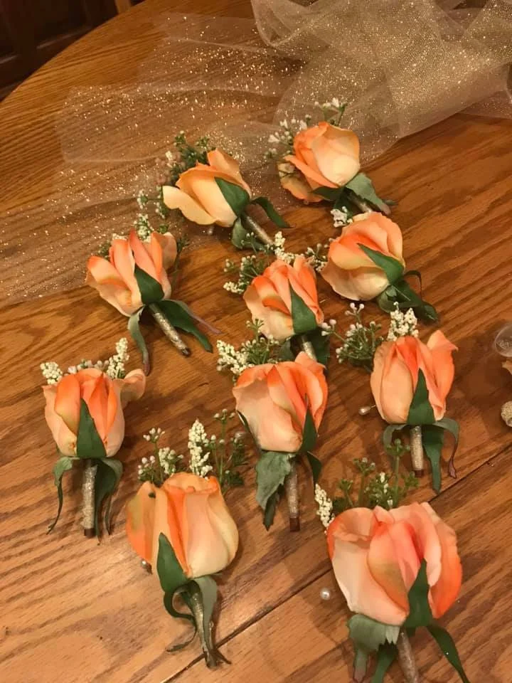A collection of peach-colored rose boutonnieres with green leaves and small white filler flowers arranged on a wooden surface.