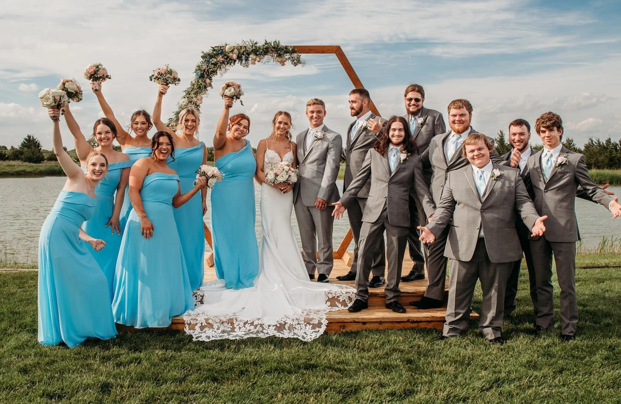 Wedding party outdoors by a lake with celebrating people, women in blue dresses, men in gray suits, a bride in white gown, all smiling, holding bouquets, standing on wooden platform under a floral arch.