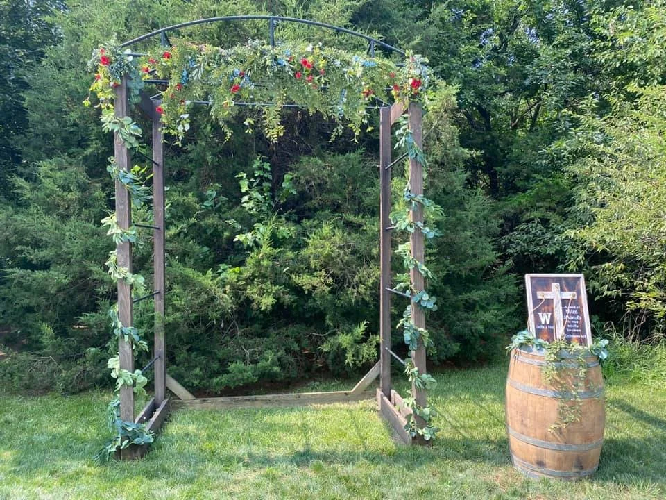 Decorative outdoor arch with flowers and greenery, next to a barrel with a framed sign featuring a cross and text, set in a grassy area with trees in the background.