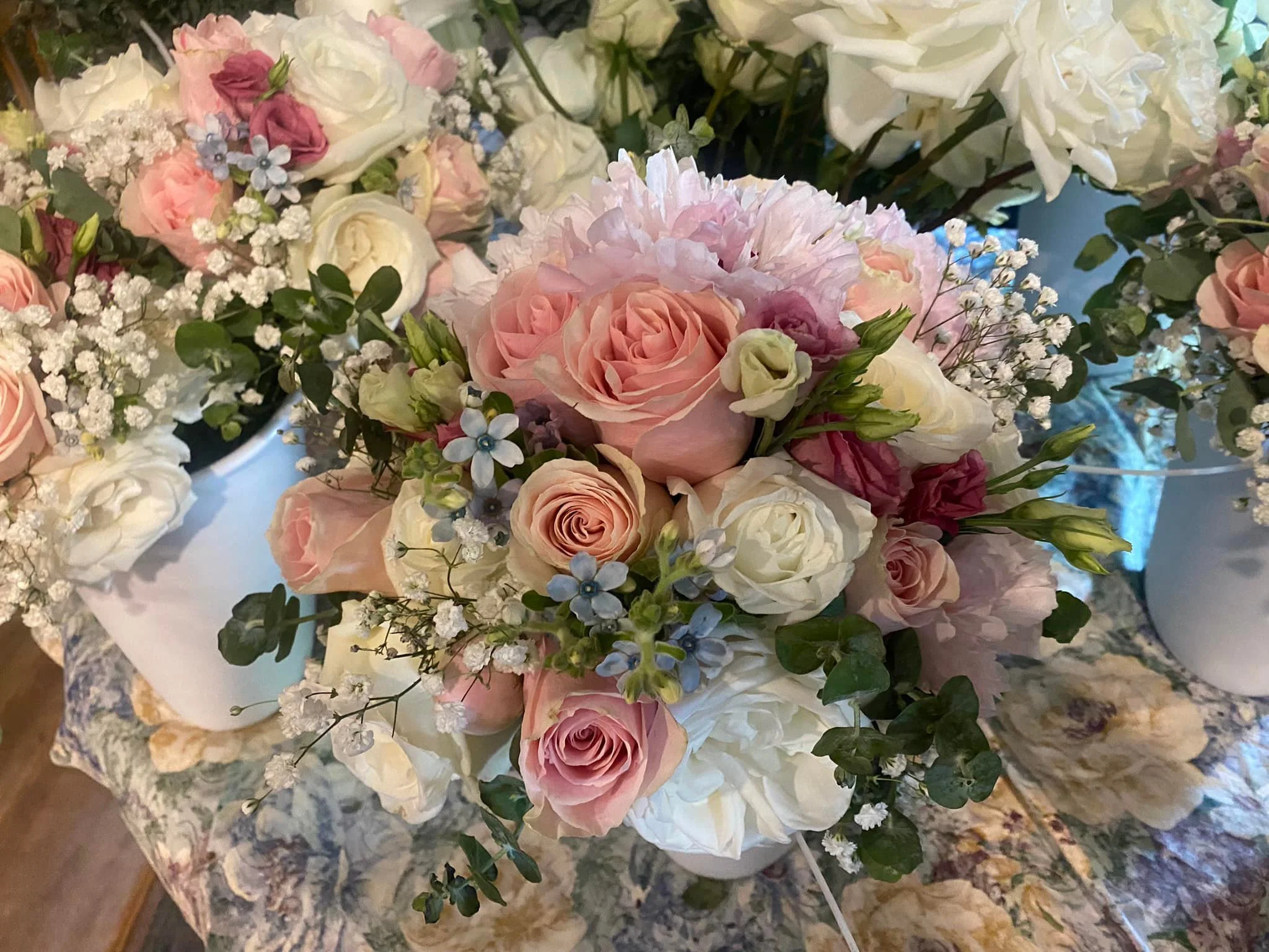 A lush bouquet of pastel pink, white, and light purple roses, along with small white and blue flowers and green foliage, arranged in a white vase on a floral-patterned tablecloth.