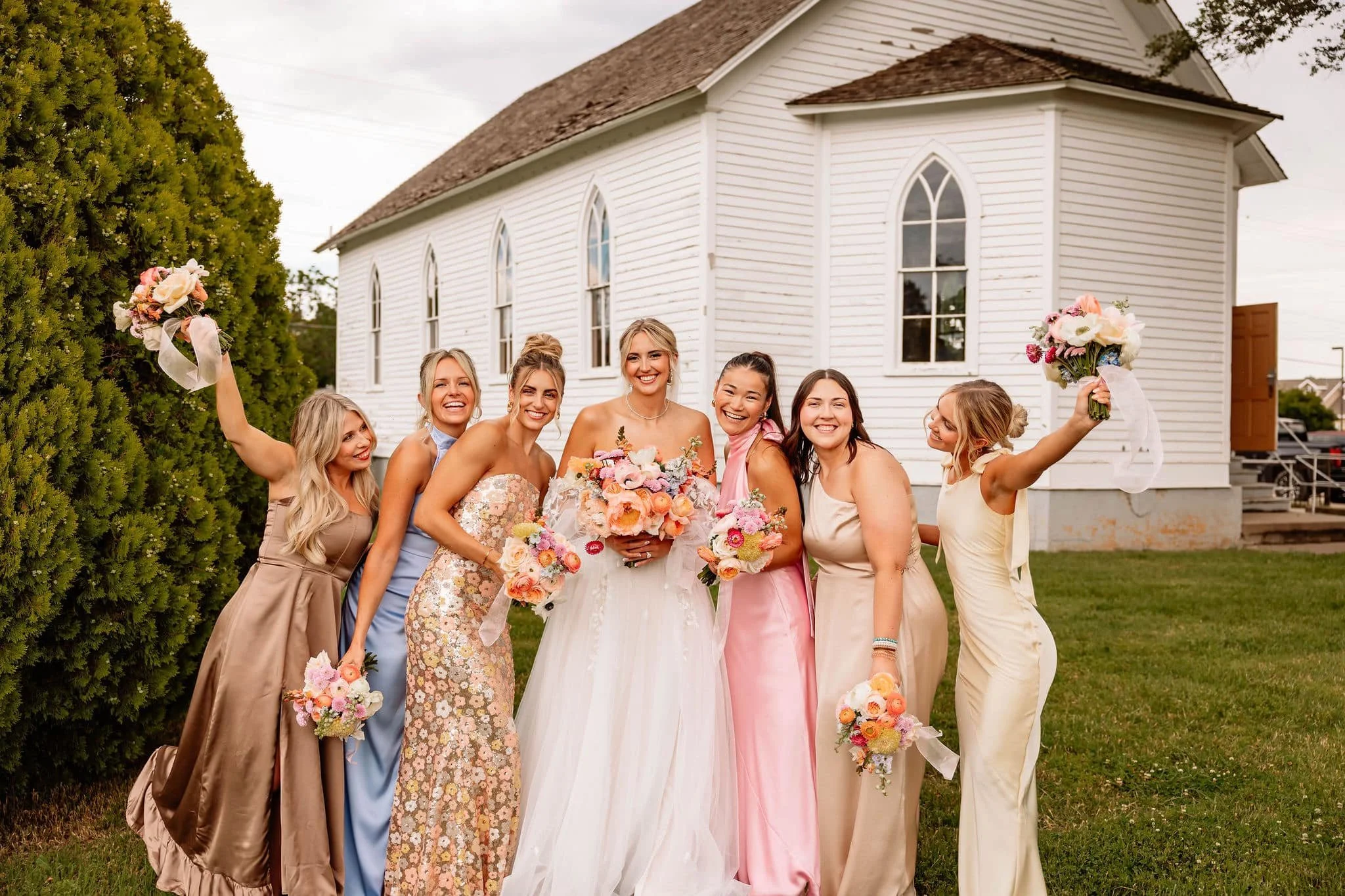 Group of women in dresses standing outside a white church, smiling, holding bouquets of flowers, during a wedding celebration.