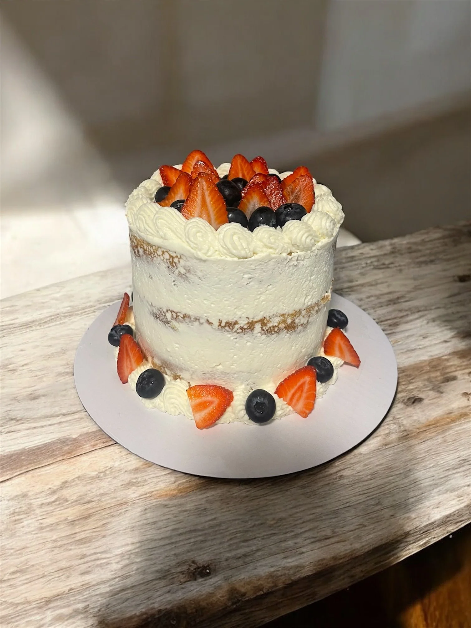 A multi-layered white frosted cake decorated with sliced strawberries and blueberries on top and around the base, placed on a white circular cake board on a rustic wooden surface.