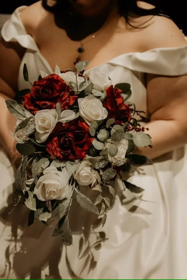 A woman in a white off-shoulder dress holding a bouquet of white and red roses with eucalyptus leaves.