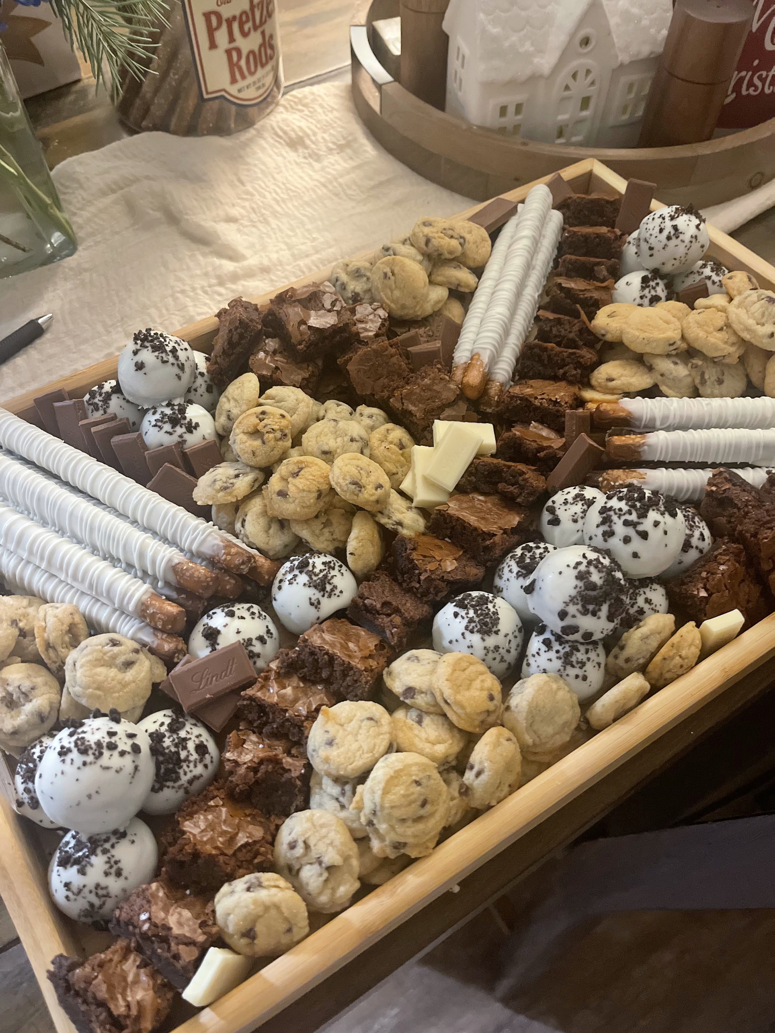 Assorted holiday treats on a wooden tray, including chocolate brownies, chocolate chip cookies, white chocolate sticks, white chocolate with sprinkles, and chocolate with sprinkles, with a festive setting and candles in the background.