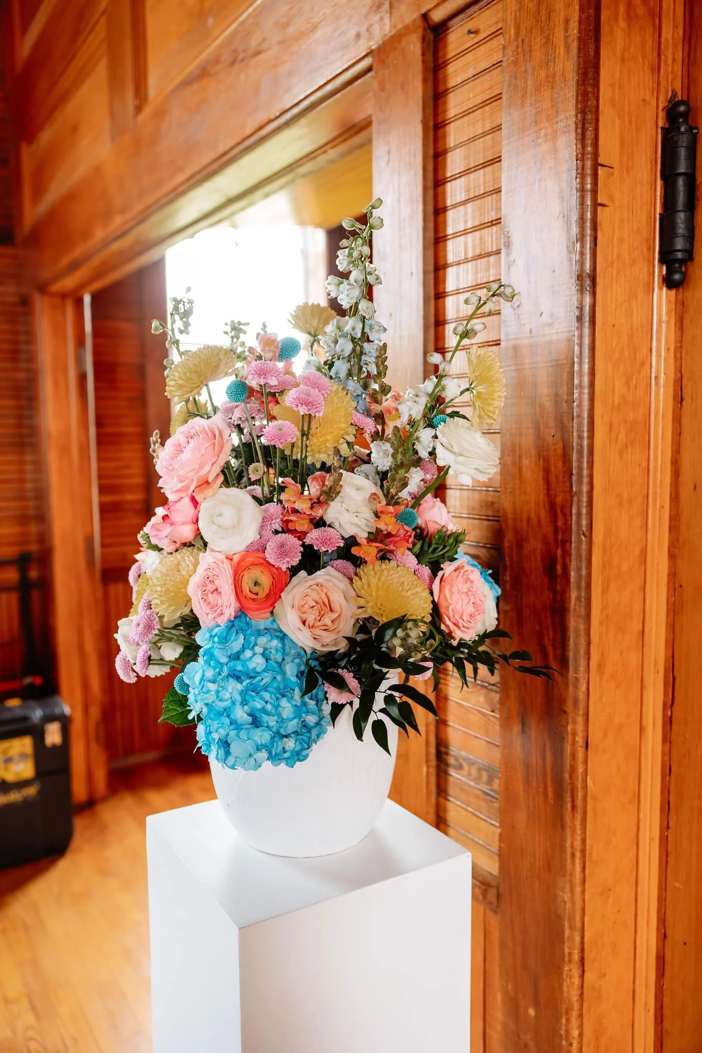Colorful flower arrangement in a white vase on a white pedestal, placed against a wood-paneled wall.