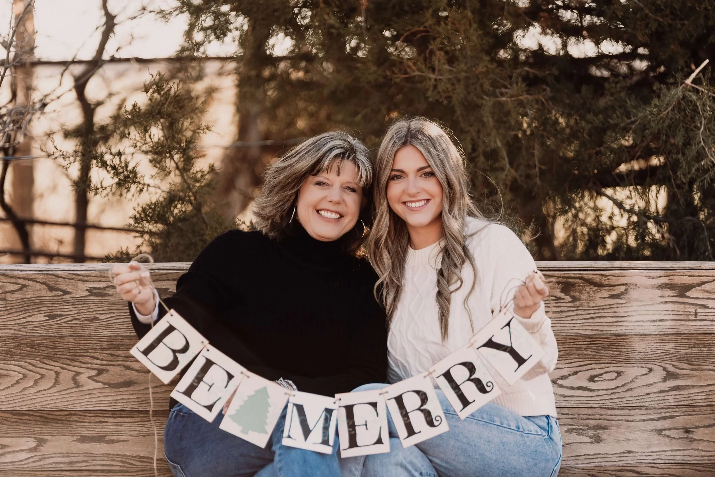 Two smiling women sitting on a wooden bench outdoors holding a 'Best Mom' banner. The woman on the left is wearing a black sweater, and the woman on the right is wearing a white sweater. There are trees in the background.