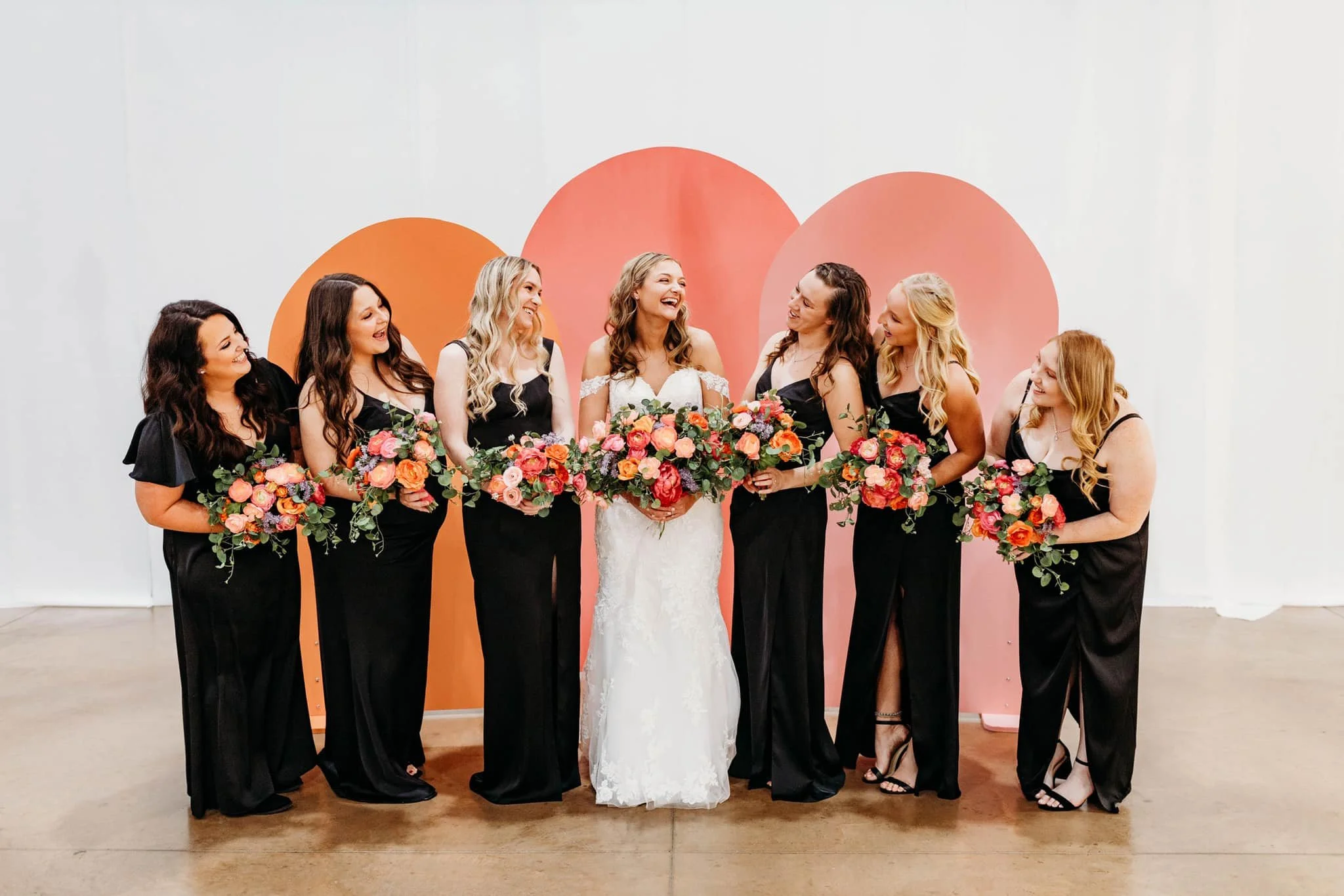 A bride in a white wedding dress holding a bouquet of pink and red flowers, standing with six bridesmaids dressed in black, each holding a similar bouquet, in front of a decorative backdrop of large pink and orange circles.
