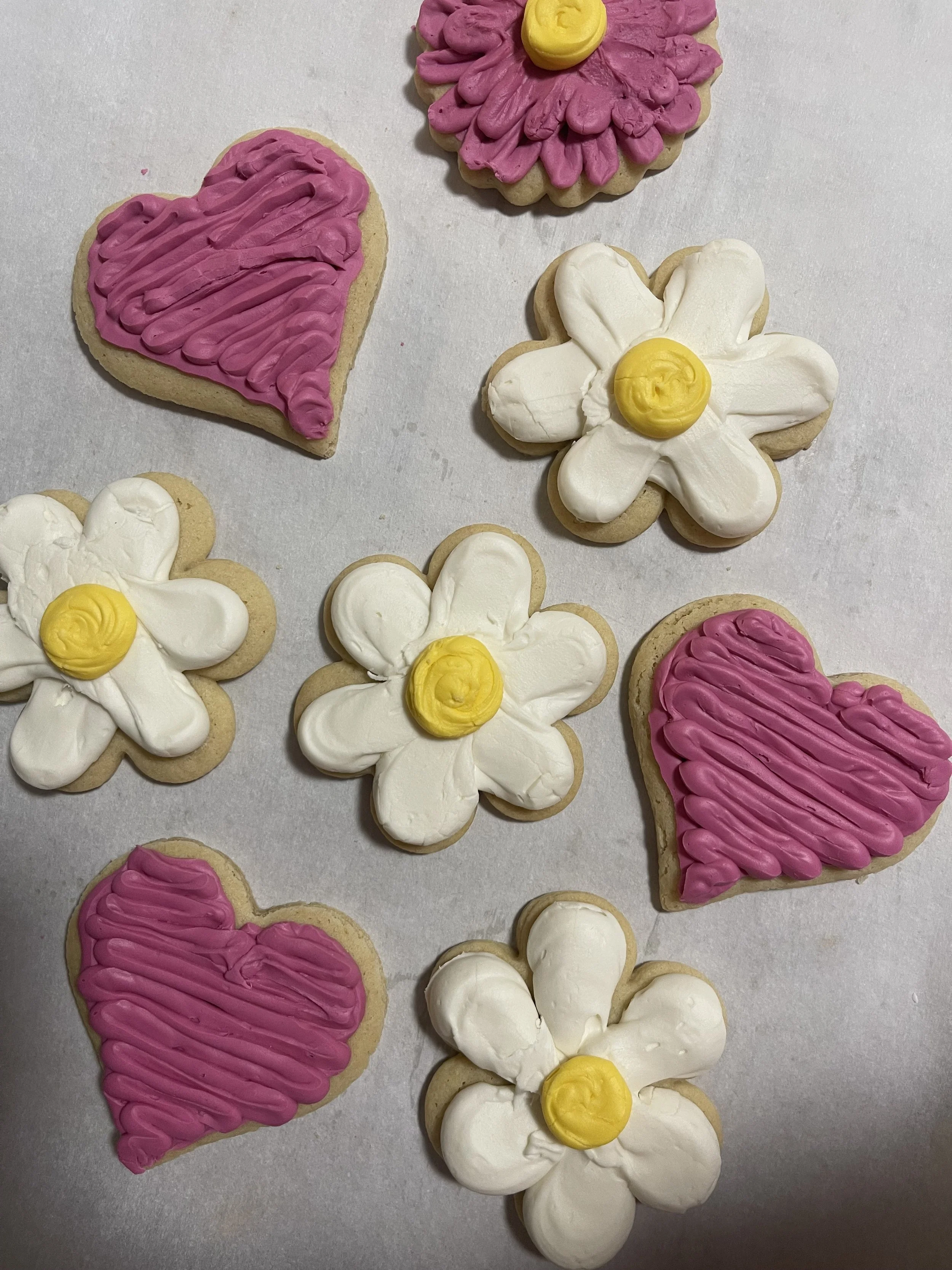 Decorated cookies with pink hearts, white daisies with yellow centers, arranged on white parchment paper.