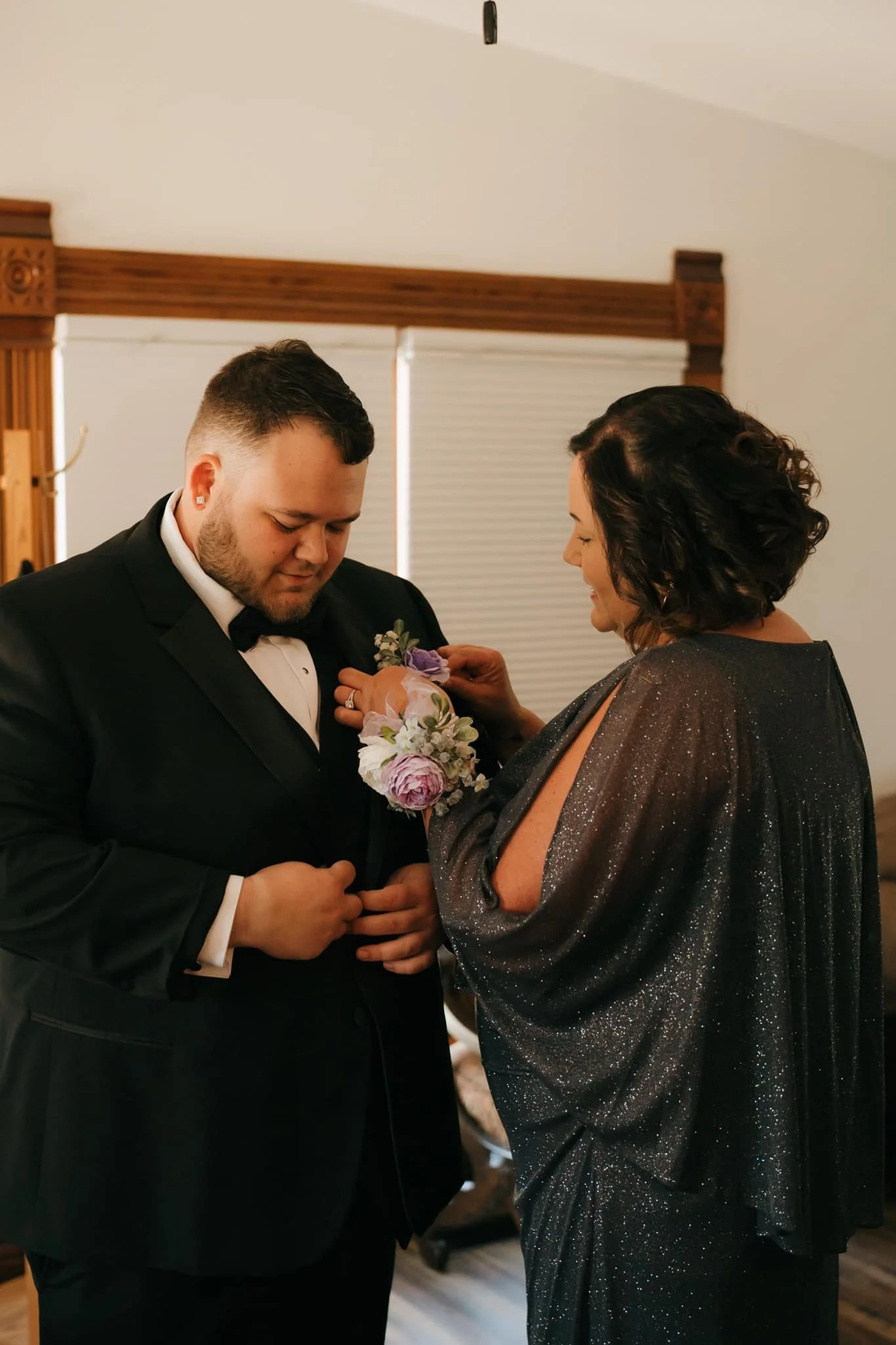 The groom in a tuxedo being pinned with a corsage by his mom in a sparkly dress.
