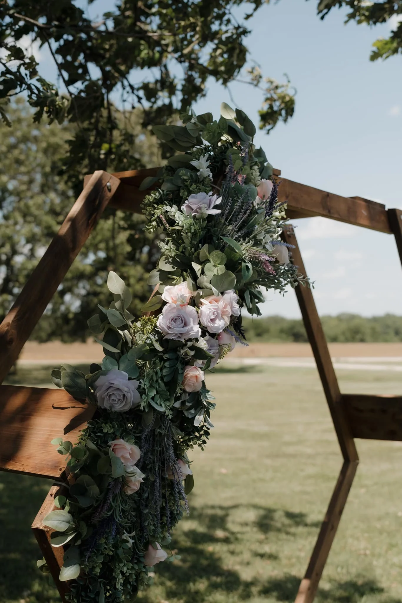 Wooden arch decorated with a cascading floral arrangement of roses, greenery, and lavender, set outdoors near a body of water, under a partially cloudy sky.