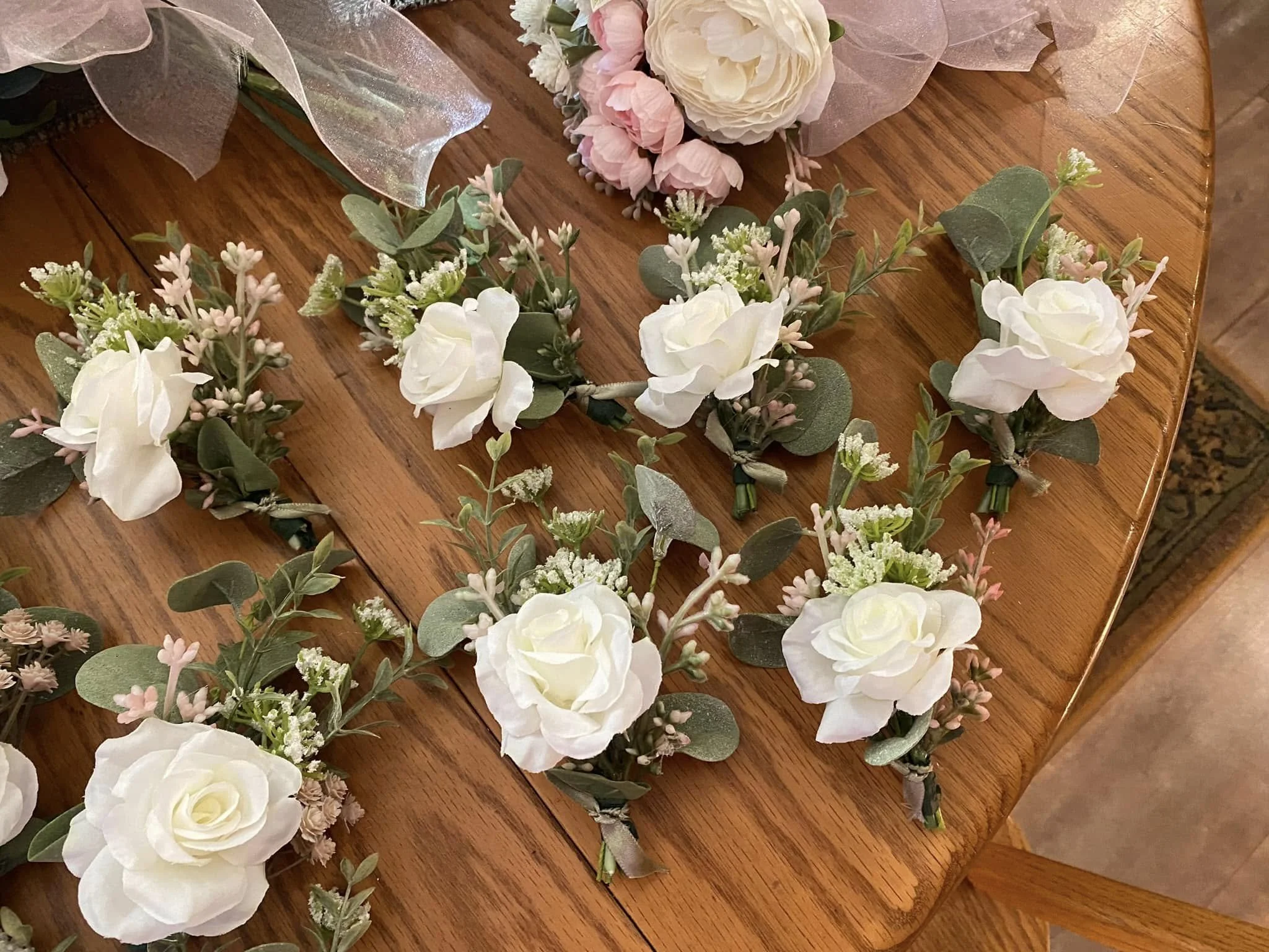 Various white and pink floral corsages with greenery and small flowers on a wooden table.