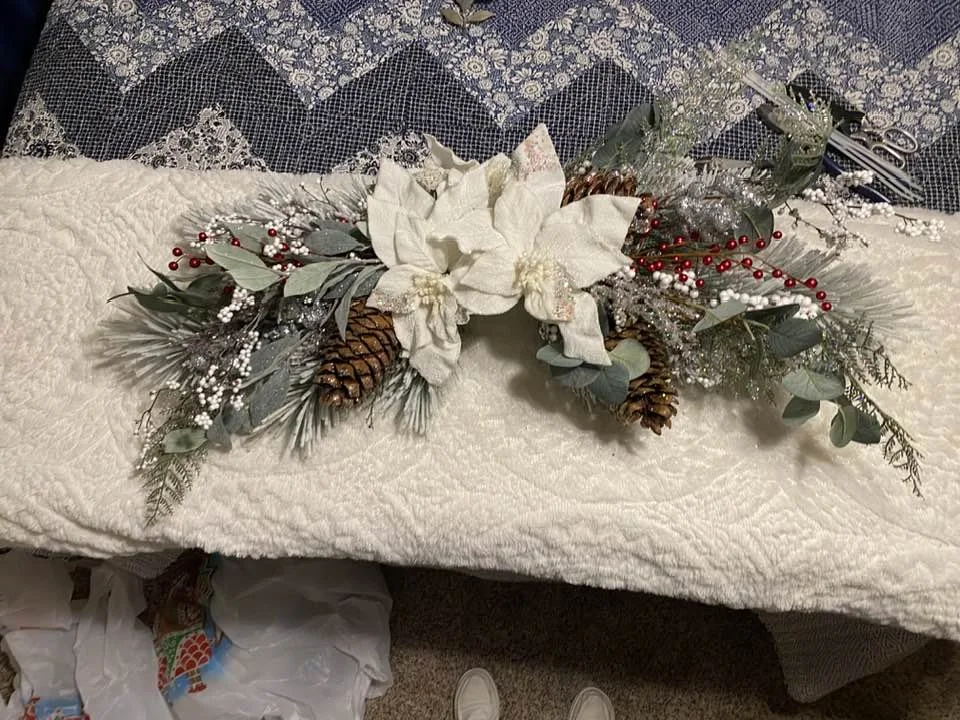 A holiday centerpiece arrangement with white poinsettias, pinecones, red berries, silver and white decorative elements, and greenery, placed on a cream-colored textured fabric.
