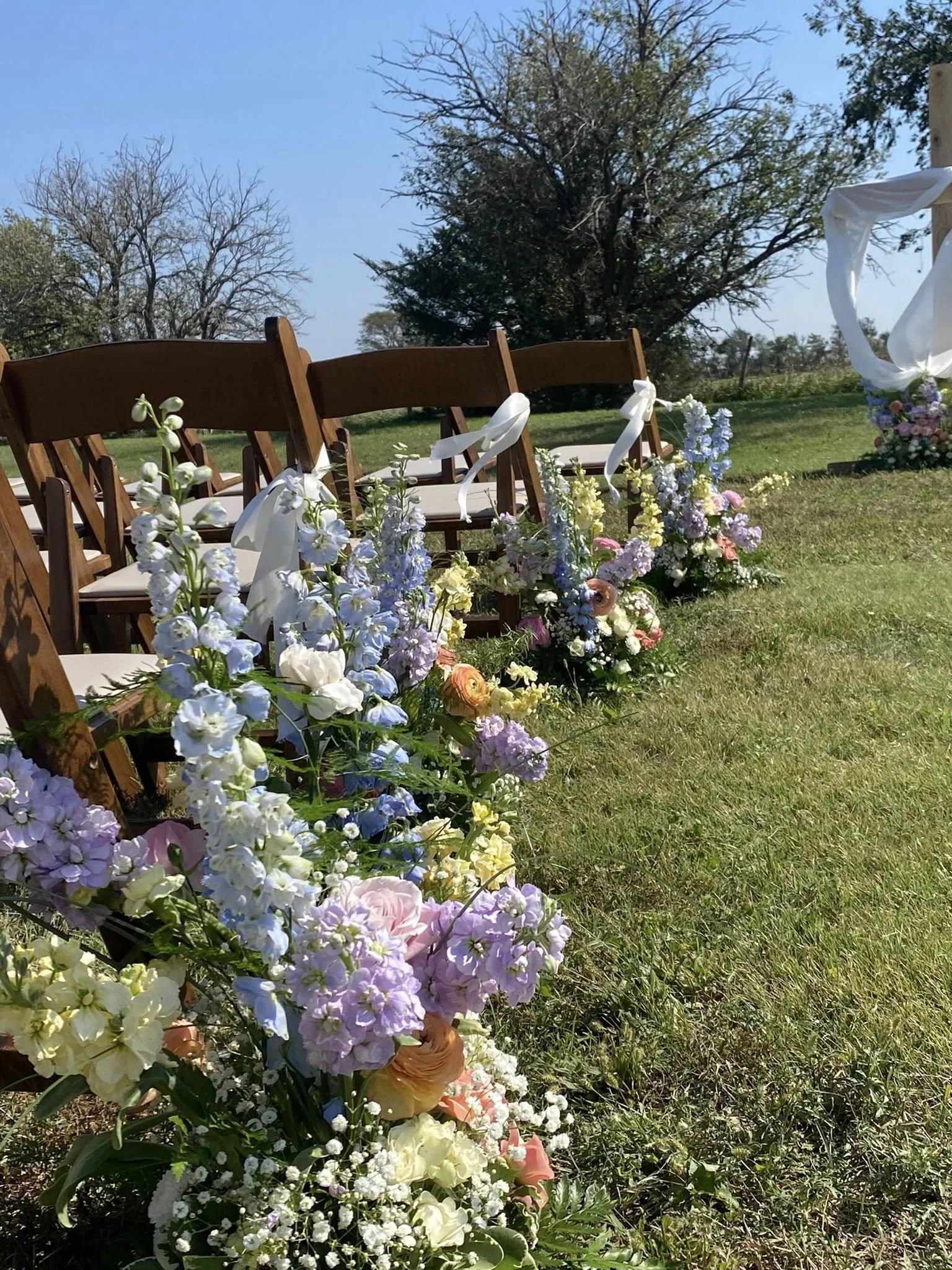 Outdoor wedding setup with wooden chairs decorated with white ribbons arranged in a row, decorated with colorful flowers including roses, delphiniums, and baby's breath along the aisle on green grass, trees in the background, under a clear blue sky.