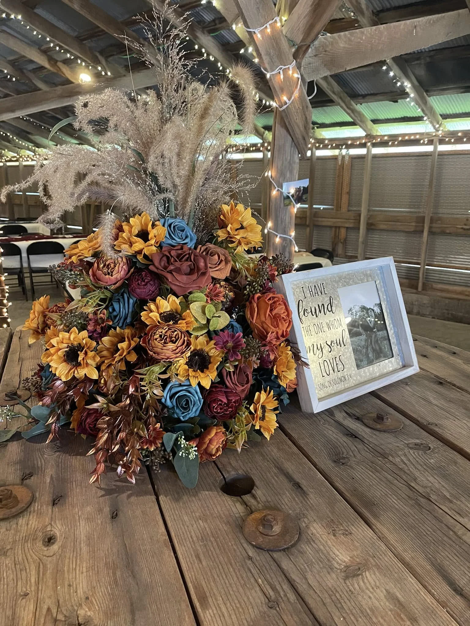 A rustic wooden table with a large bouquet of colorful flowers and a framed quote that reads, "I have found the one whom my soul loves," decorated with string lights in a barn setting.