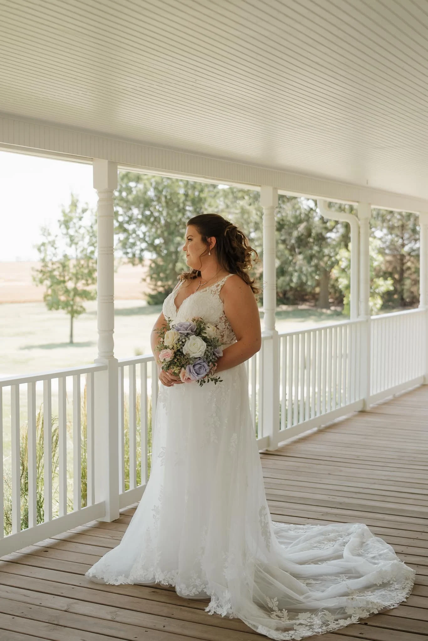 A bride in a white wedding gown holding a bouquet of colorful flowers on a porch with white railings, overlooking a green yard and trees.
