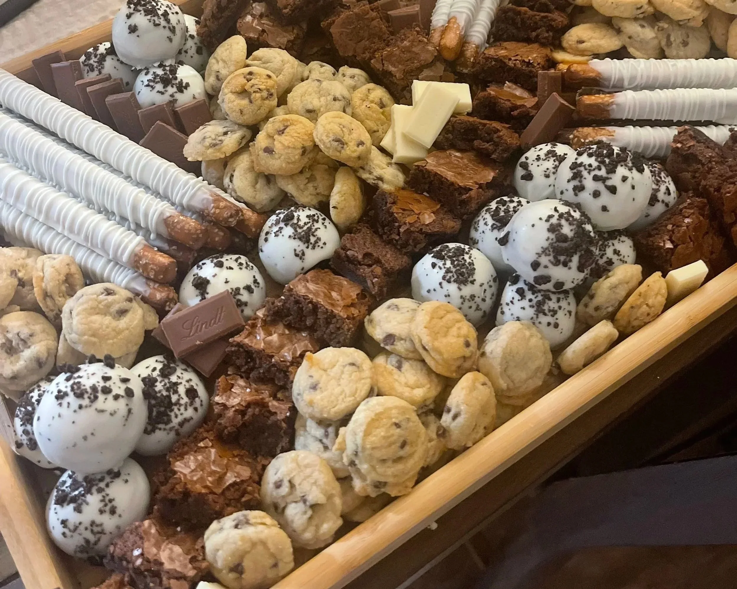 Assorted cookies, chocolate bars, white chocolate-covered pretzel rods, and decorative confections arranged on a wooden tray.