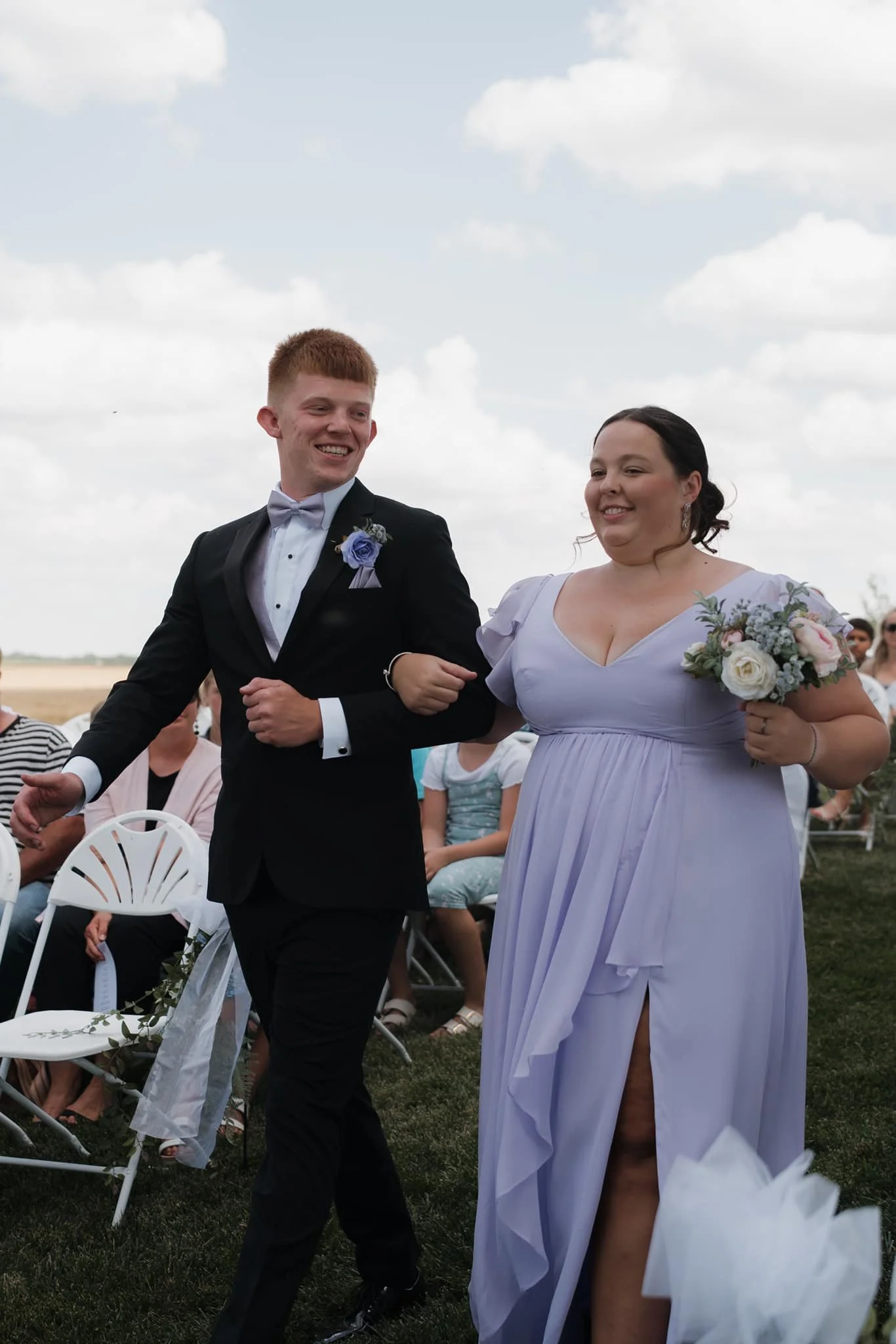 A joyful couple walking together outdoors during their wedding ceremony, with guests seated in the background.