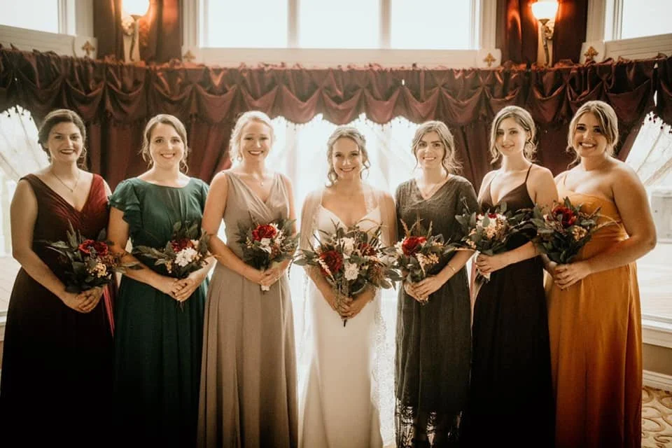 Seven women in formal dresses holding bouquets, standing in a line indoors with a window and curtains behind them.