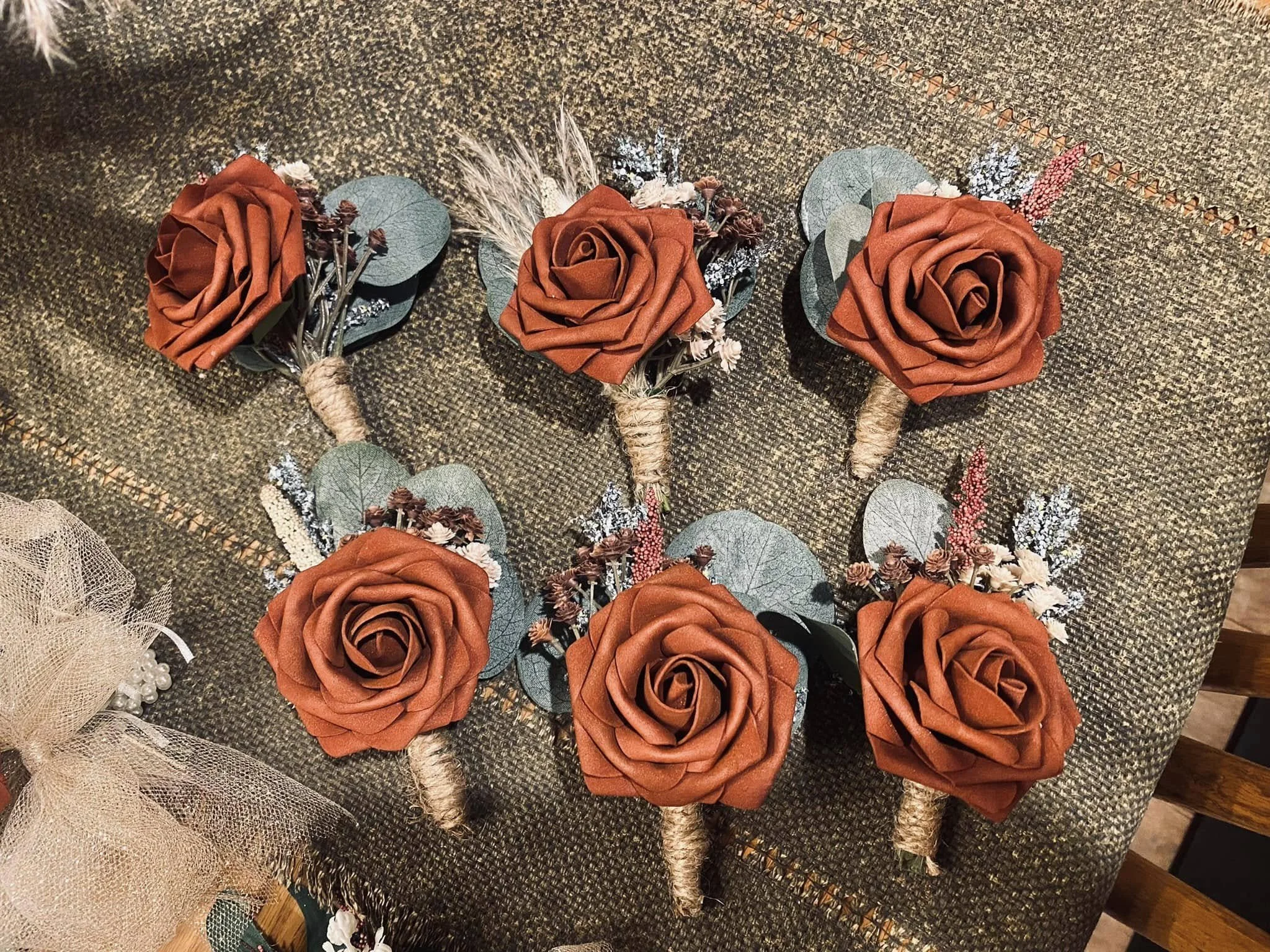Six boutonniere floral arrangements with rust-colored roses, eucalyptus leaves, small white and pink filler flowers, wrapped with twine, arranged on a textured dark fabric surface.